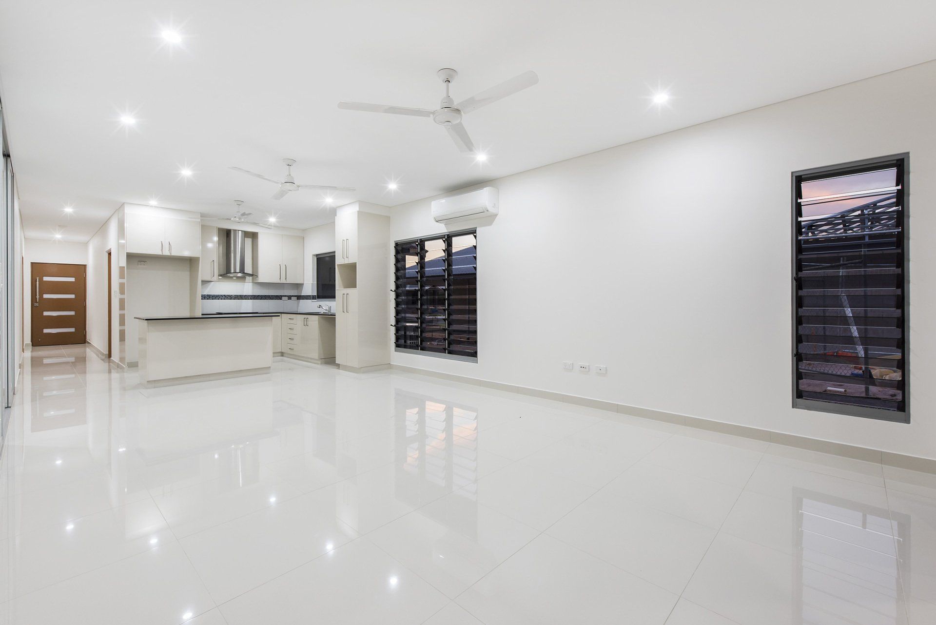 A large empty room with a ceiling fan and a kitchen in the background.
