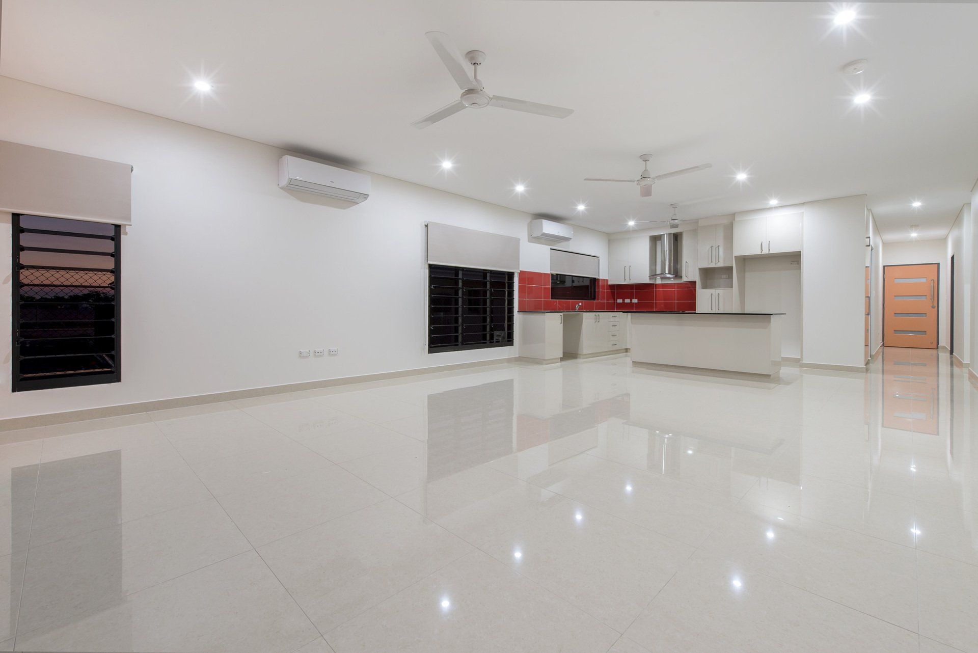 A large empty room with a ceiling fan and a kitchen in the background.