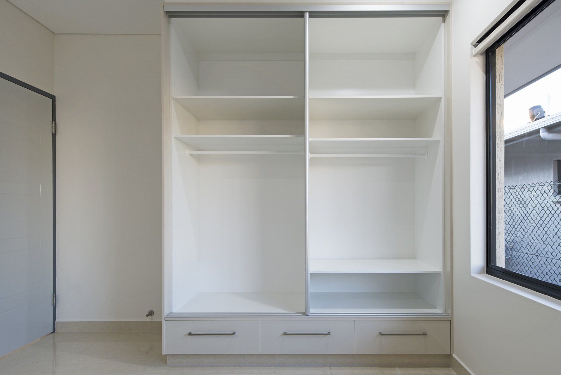An empty wardrobe with sliding doors and drawers in a bedroom with a window.