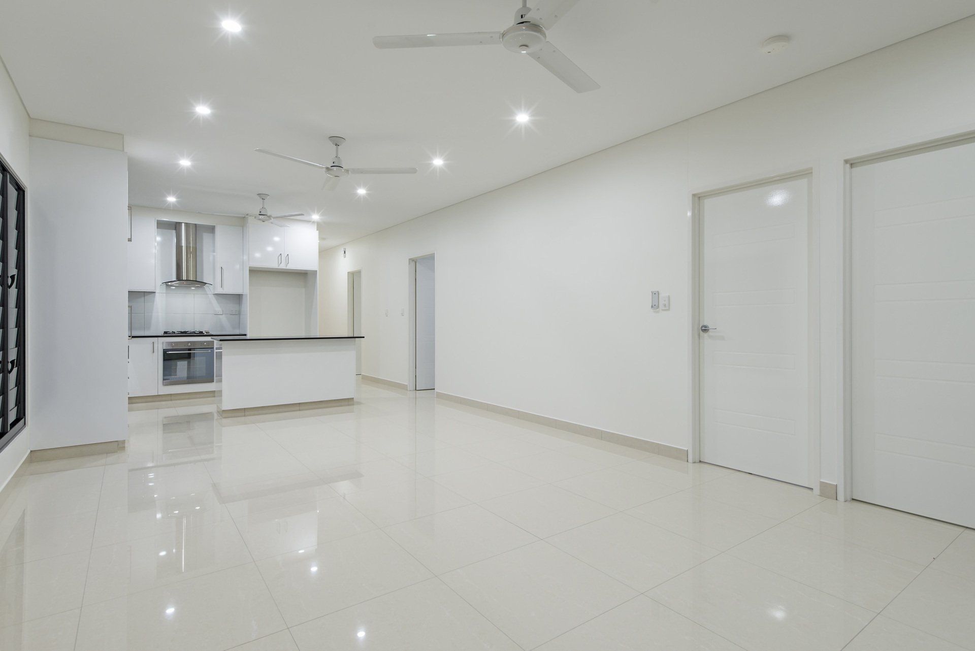 An empty living room with a ceiling fan and a kitchen in the background.