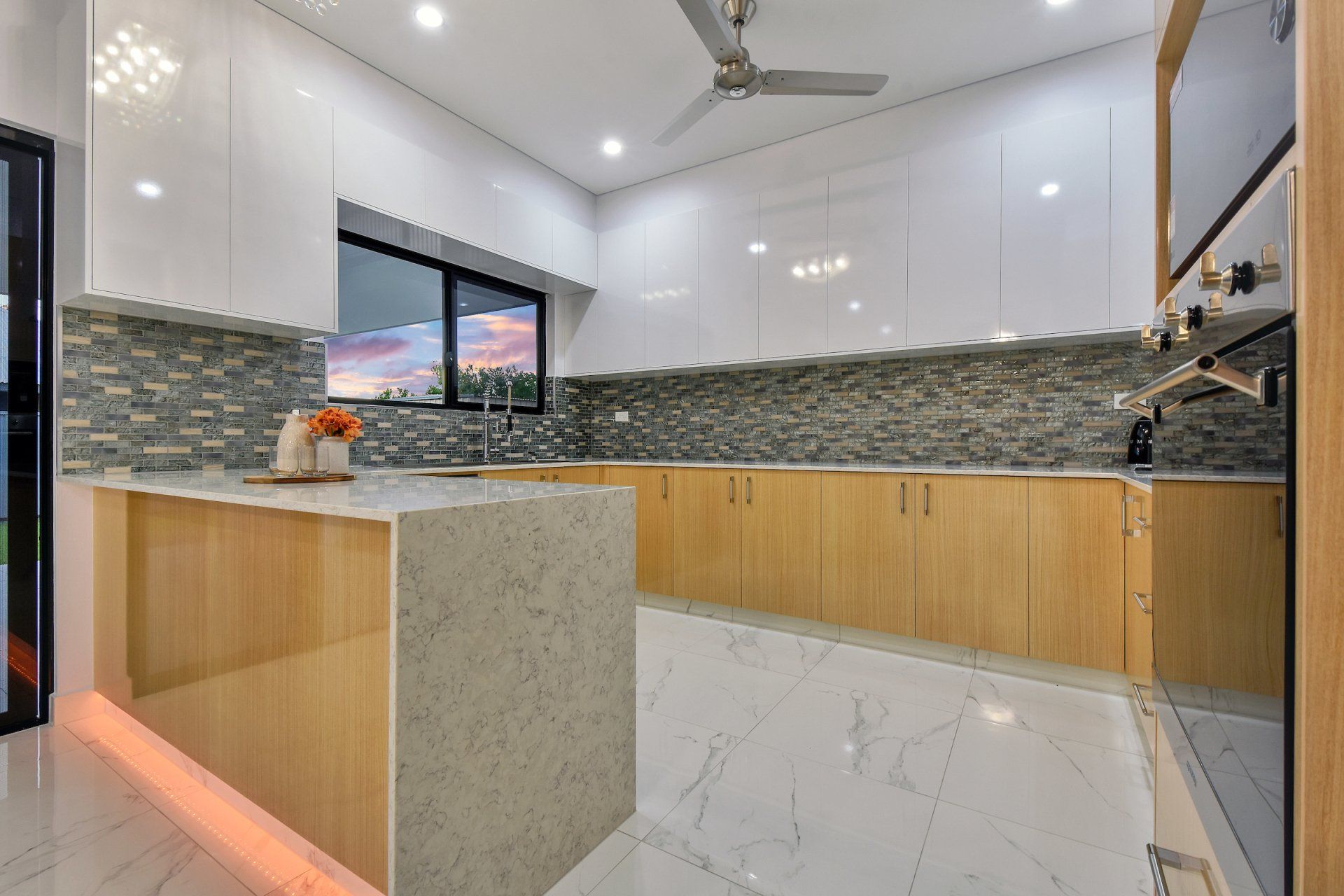 A kitchen with wooden cabinets , marble counter tops , a ceiling fan and a large island.