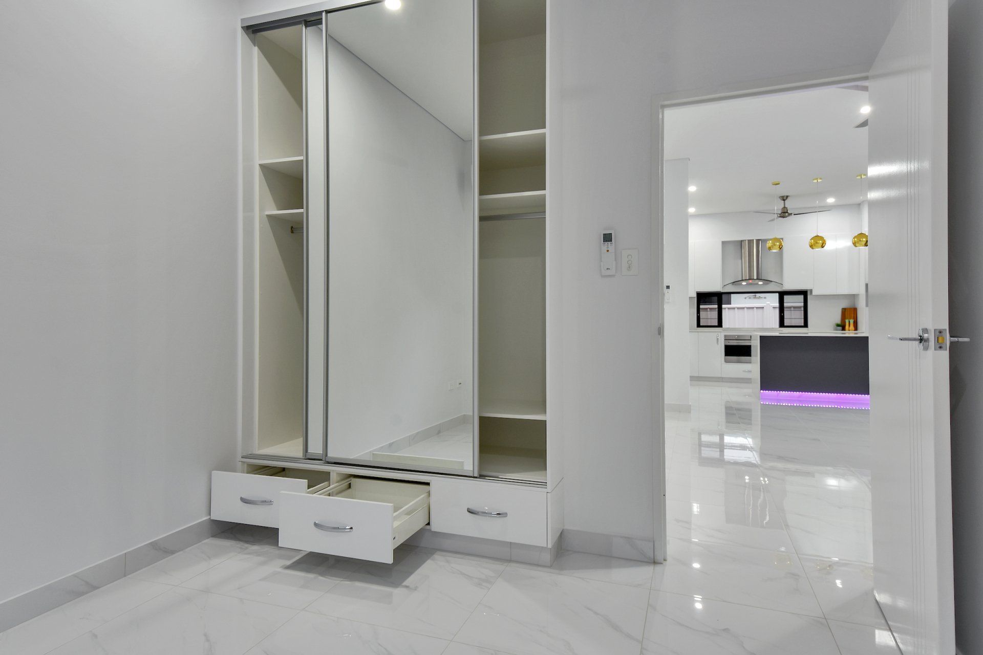 A bedroom with a large wardrobe and drawers and a kitchen in the background.