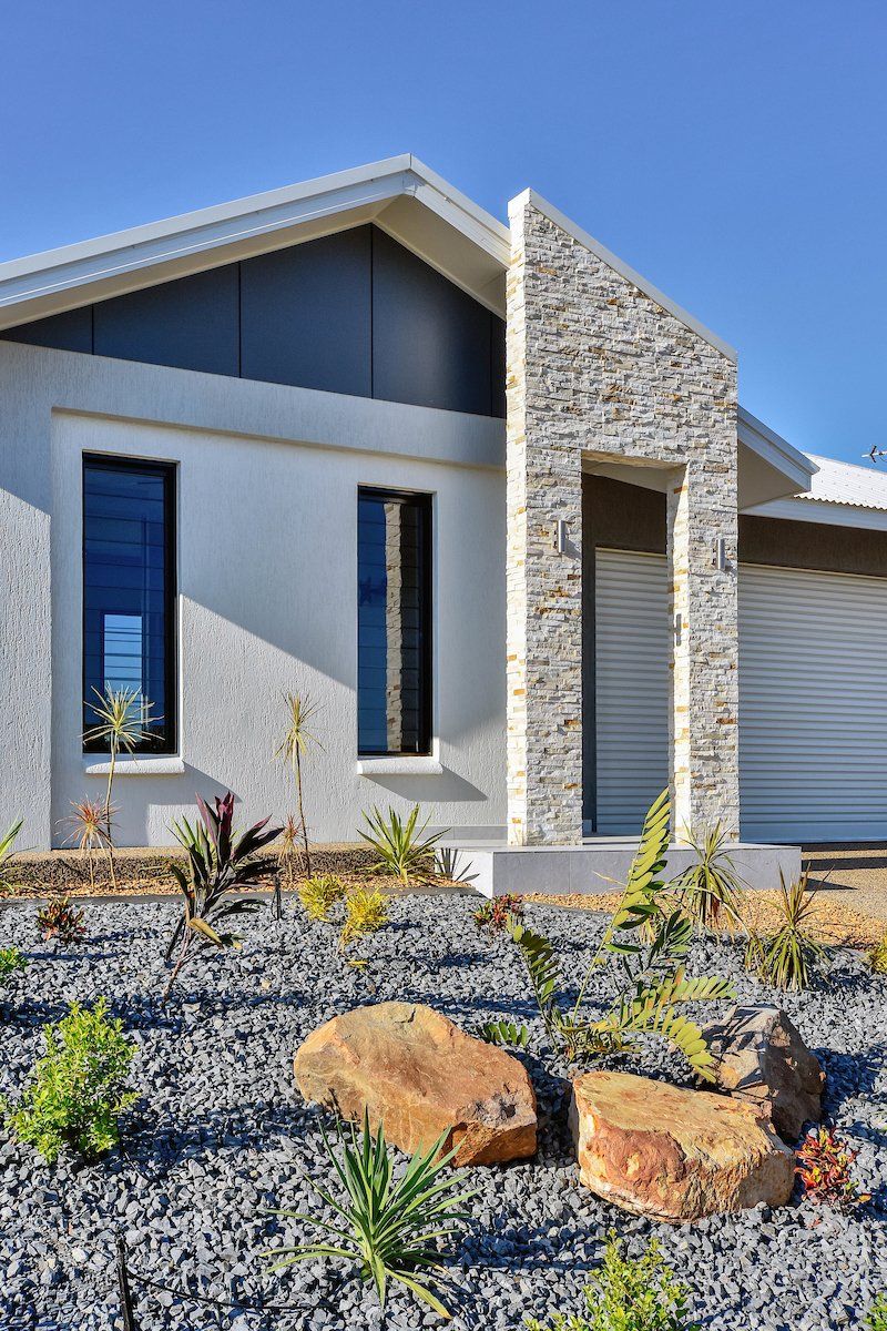 A white house with a stone facade is surrounded by rocks and plants.