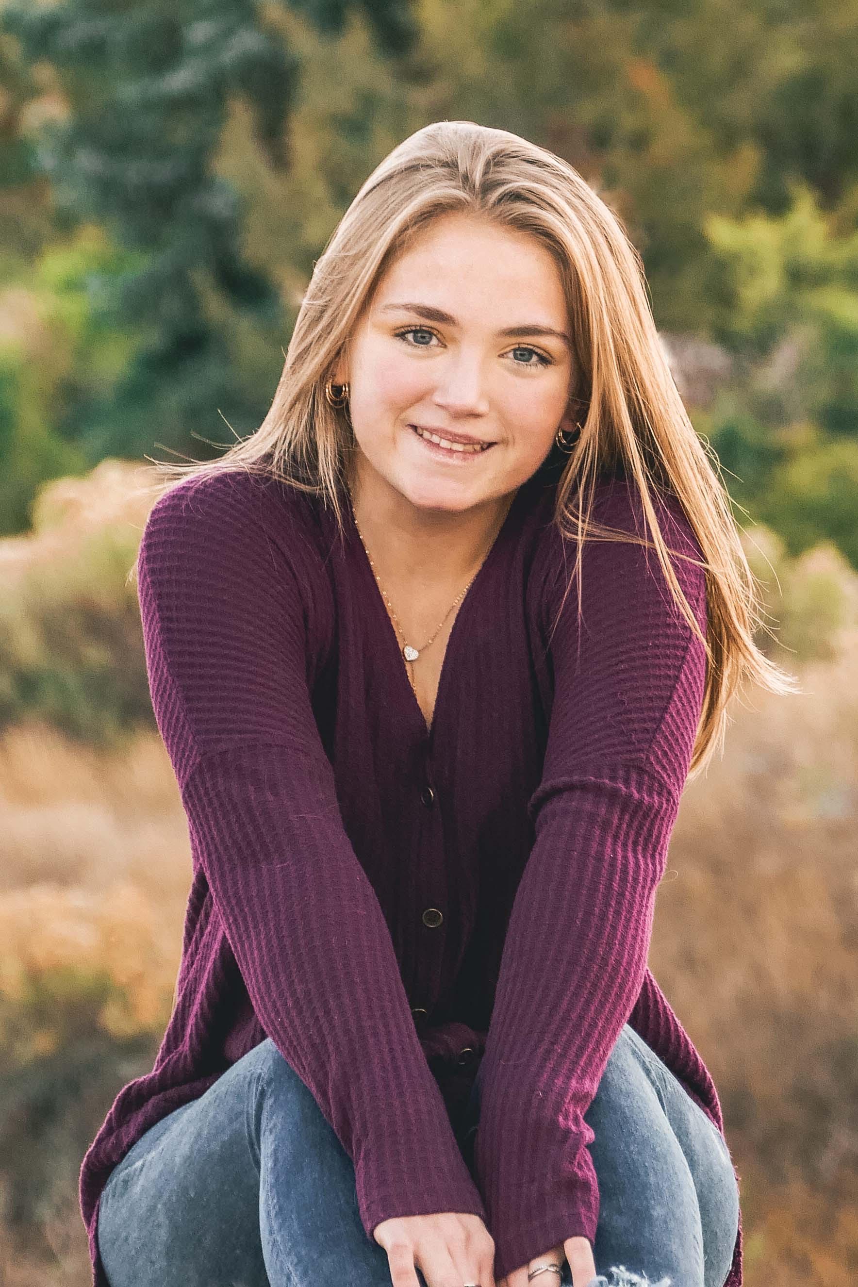A young woman in a purple sweater is kneeling down in a field.