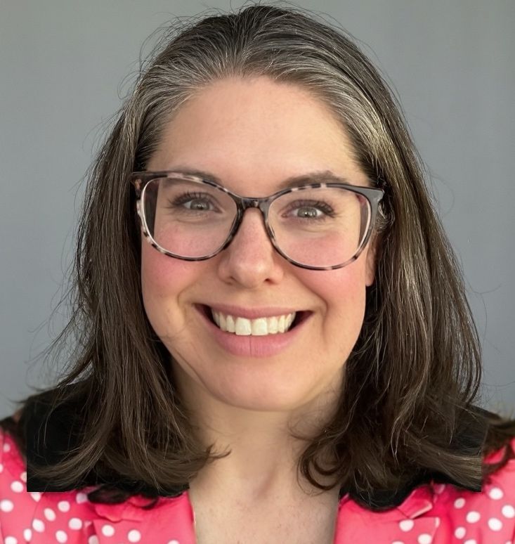 A woman wearing glasses and a pink jacket is smiling.