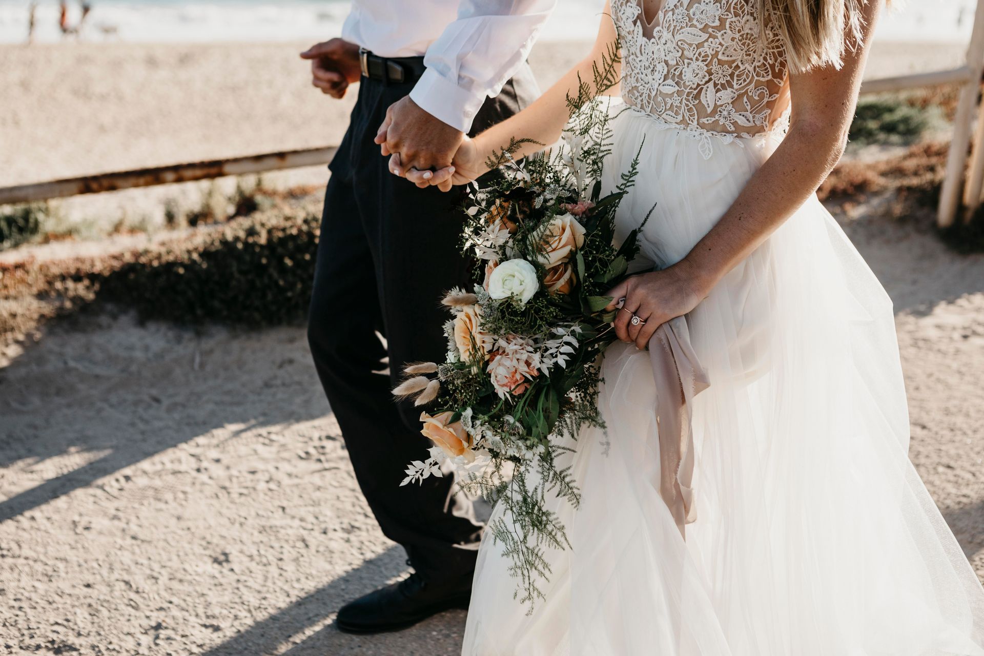 Una sposa e uno sposo si tengono per mano e camminano sulla spiaggia. La sposa tiene in mano un mazzo di fiori.