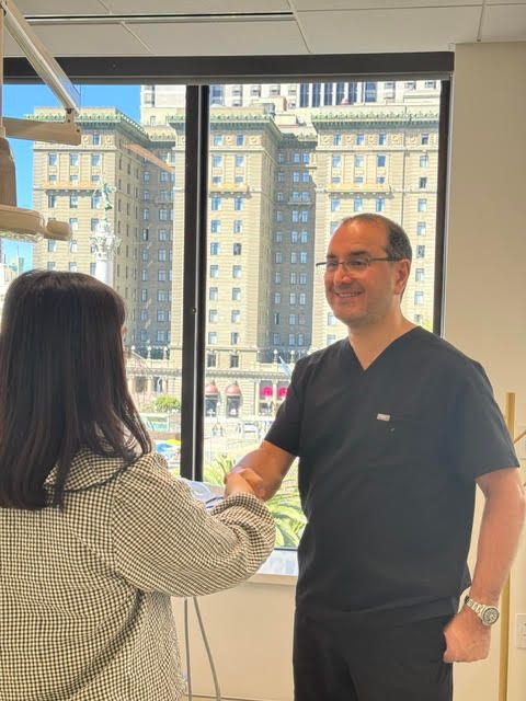 A healthcare professional in black scrubs shakes hands with a person in a patterned blazer in front of a city window.