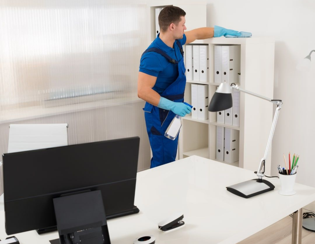 Un addetto alle pulizie professionista, in uniforme blu e guanti, pulisce una libreria in ufficio.
