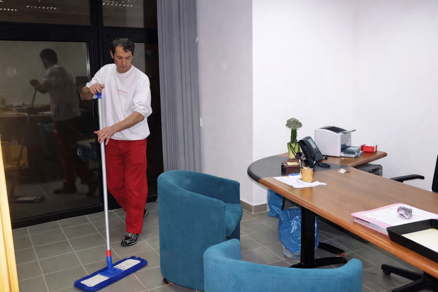 Una persona con una maglietta bianca e pantaloni rossi lava il pavimento di un ufficio arredato con poltrone blu e una scrivania di legno.