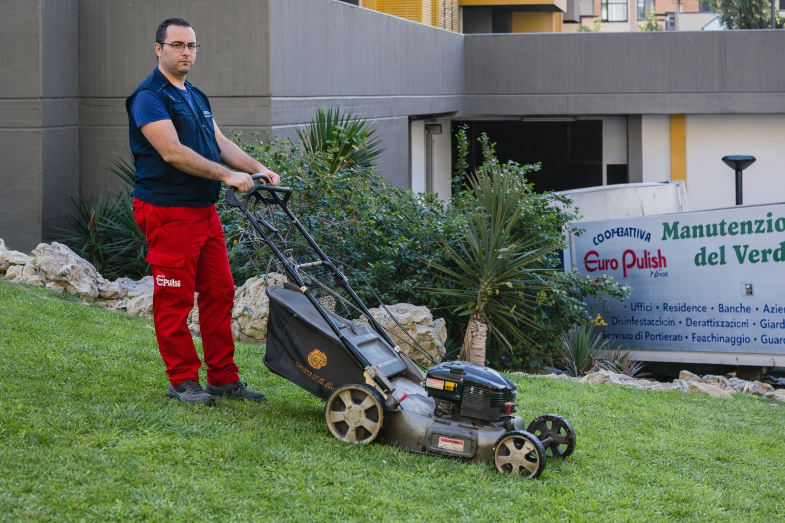 Un operaio con un gilet blu e pantaloni rossi è in piedi su un prato, intento a spingere un tosaerba davanti a un edificio con l'insegna di un'azienda.