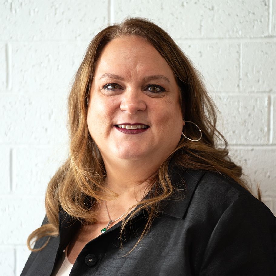 Smiling woman with blonde hair, wearing a black blazer and top, in front of a white brick wall.
