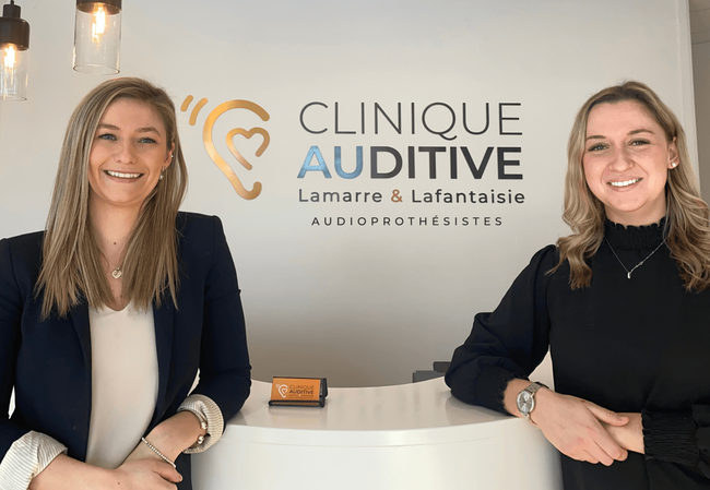 Deux audioprothésistes propriétaires se tiennent l'une à côté de l'autre devant le logo de la Clinique auditive Lamarre et Lafantaisie audioprothésistes