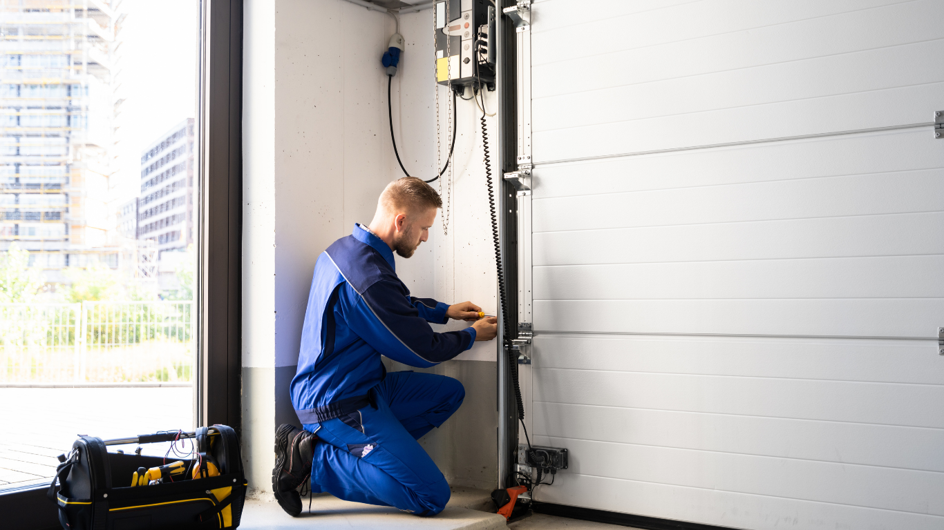 Garage door repair person in blue overalls working on a white door in a garage.