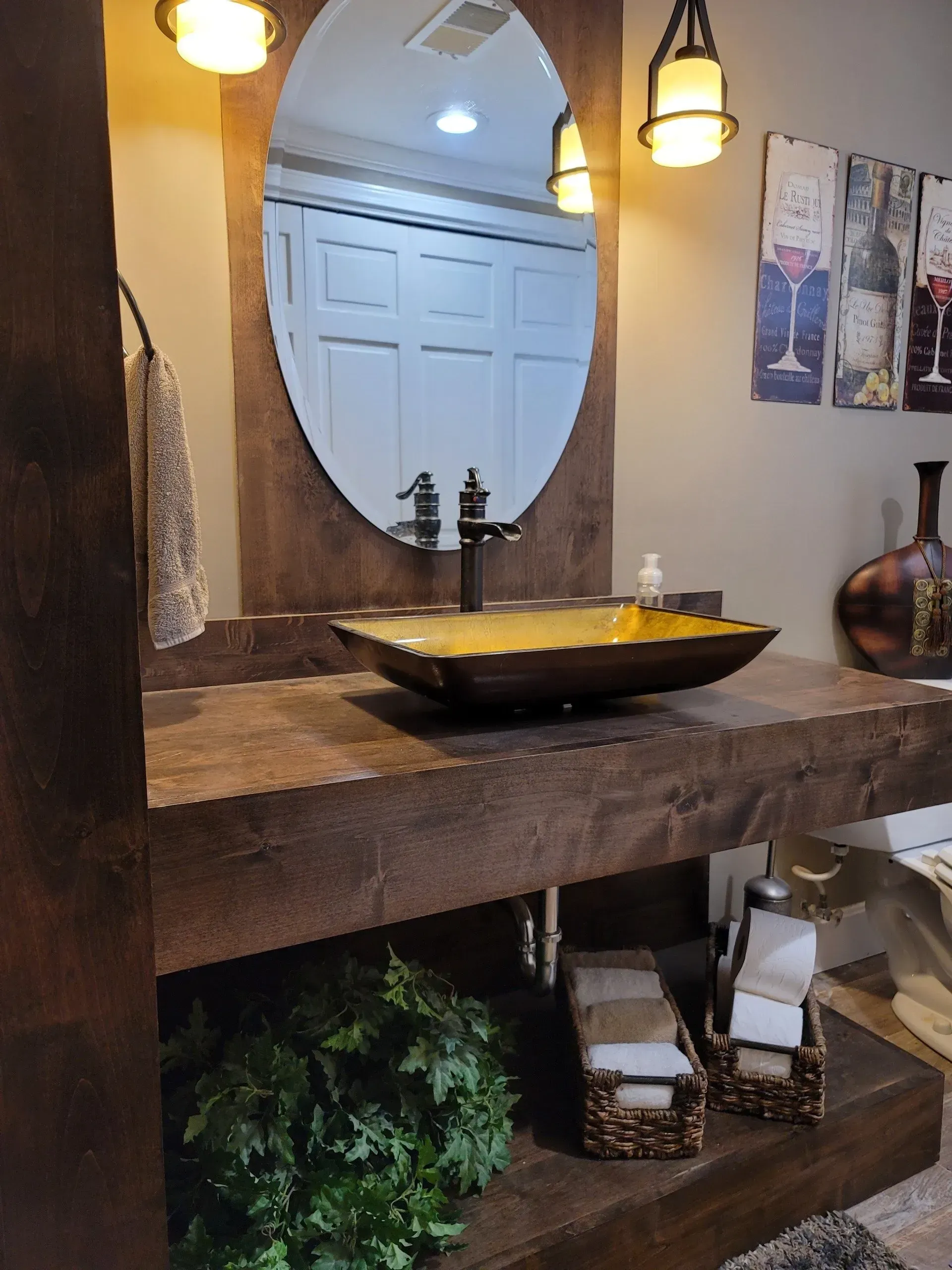 Rustic bathroom vanity with oval mirror, black sink basin, hanging lights, and decorative greenery
