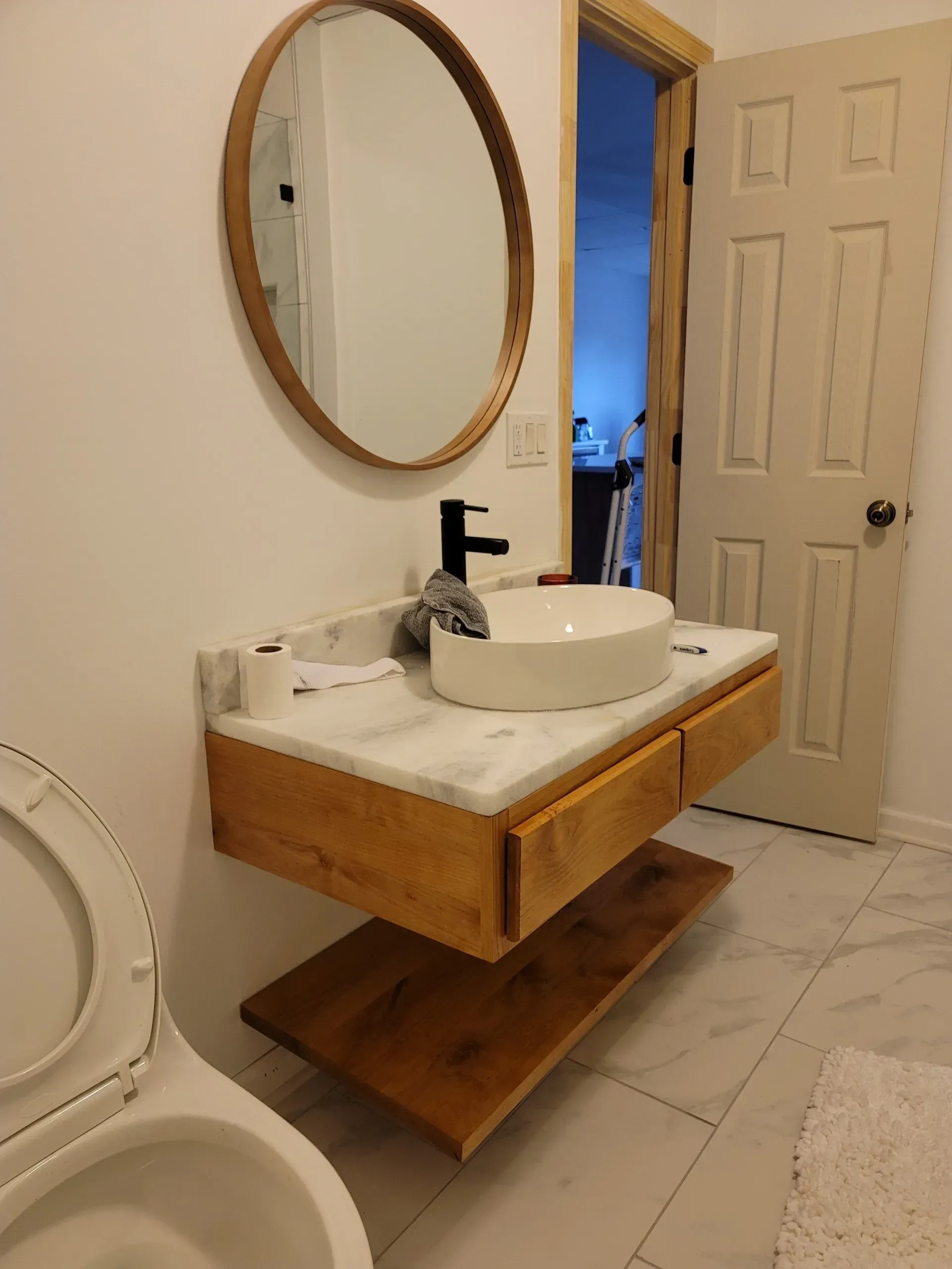 Small bathroom with wooden vanity, round mirror, white vessel sink, and open door to a blue-lit room.