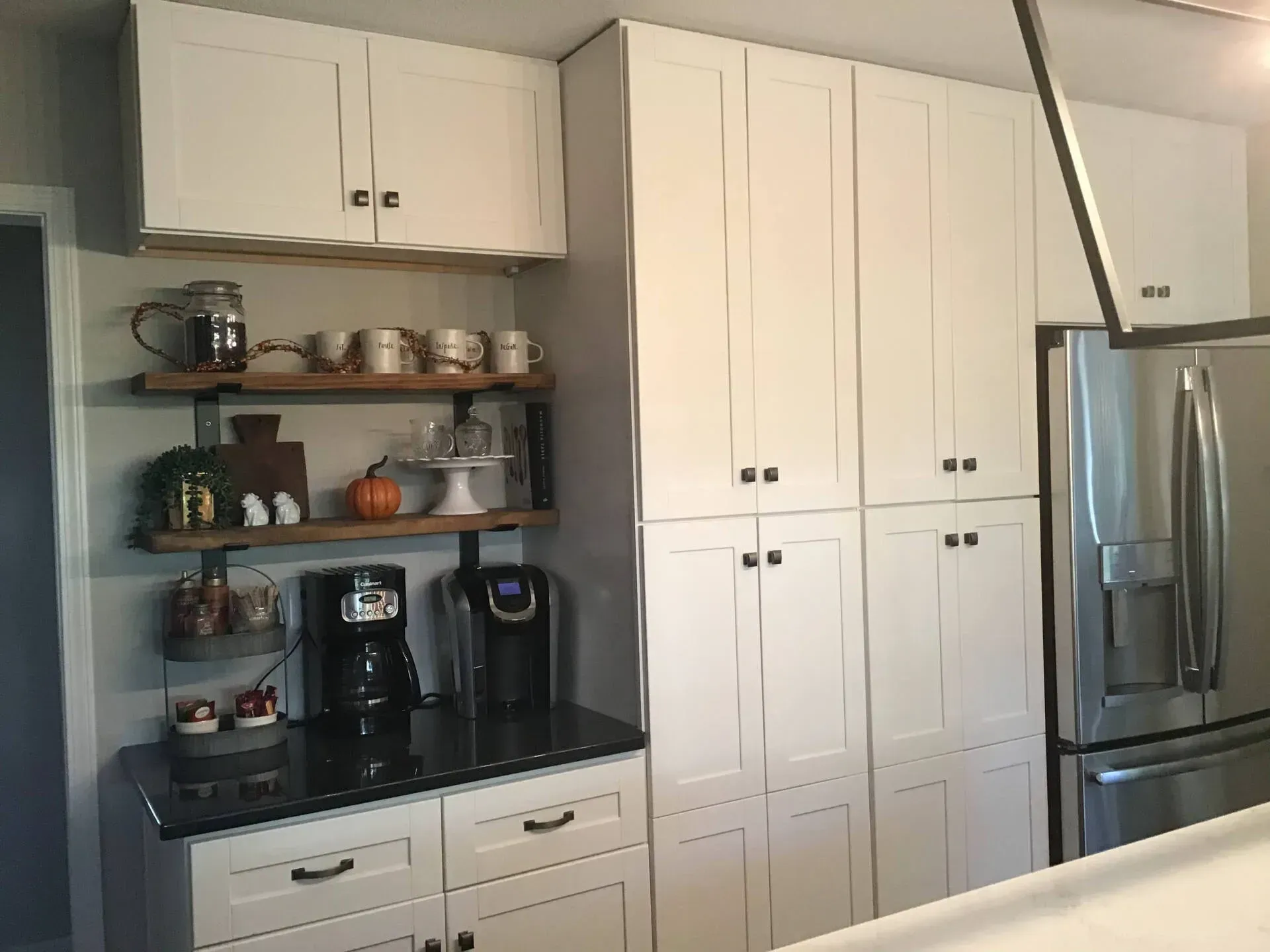 Kitchen with white cabinets, open shelves, black countertop, coffee maker, and stainless steel refrigerator.