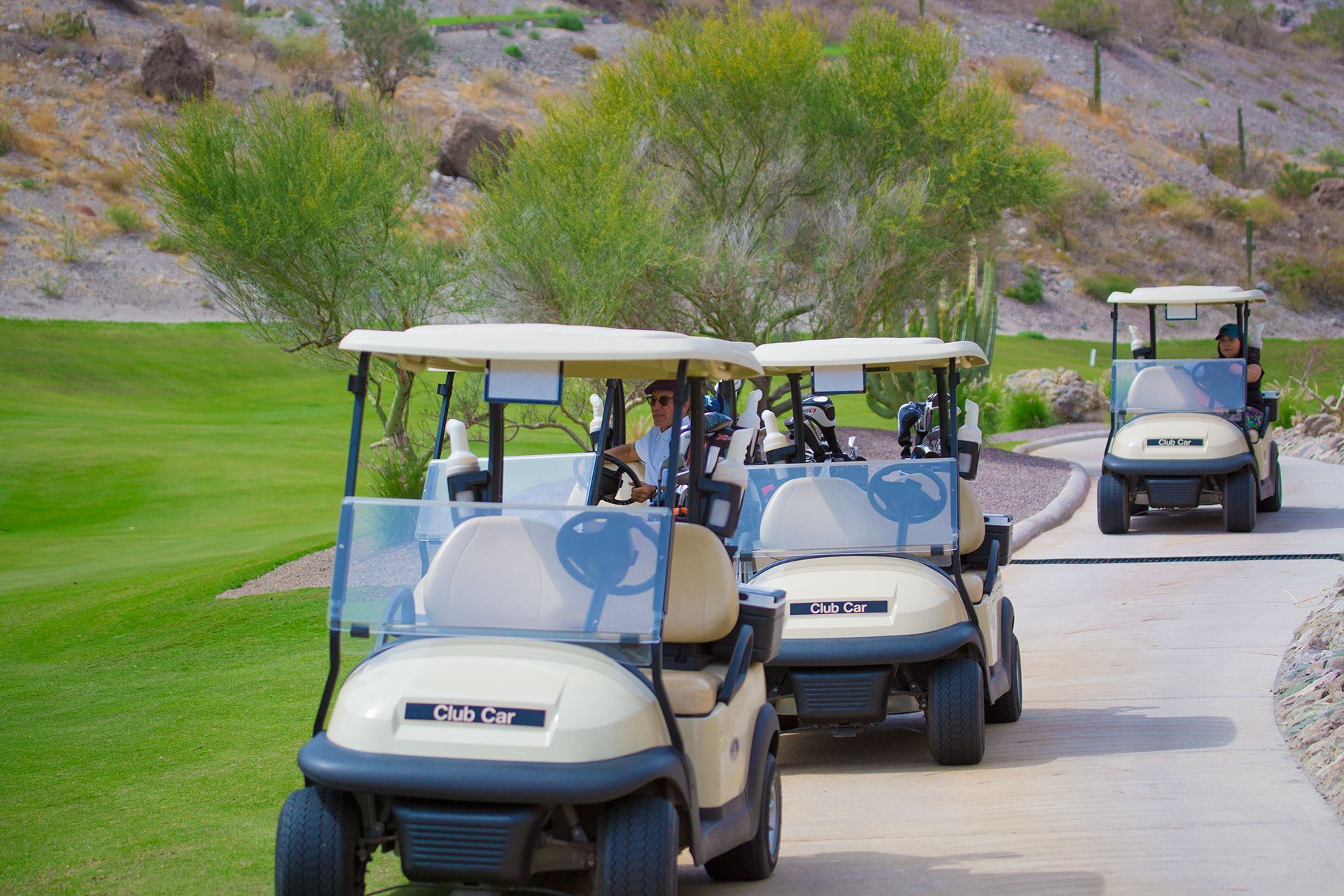 A row of golf carts are driving down a path on a golf course.