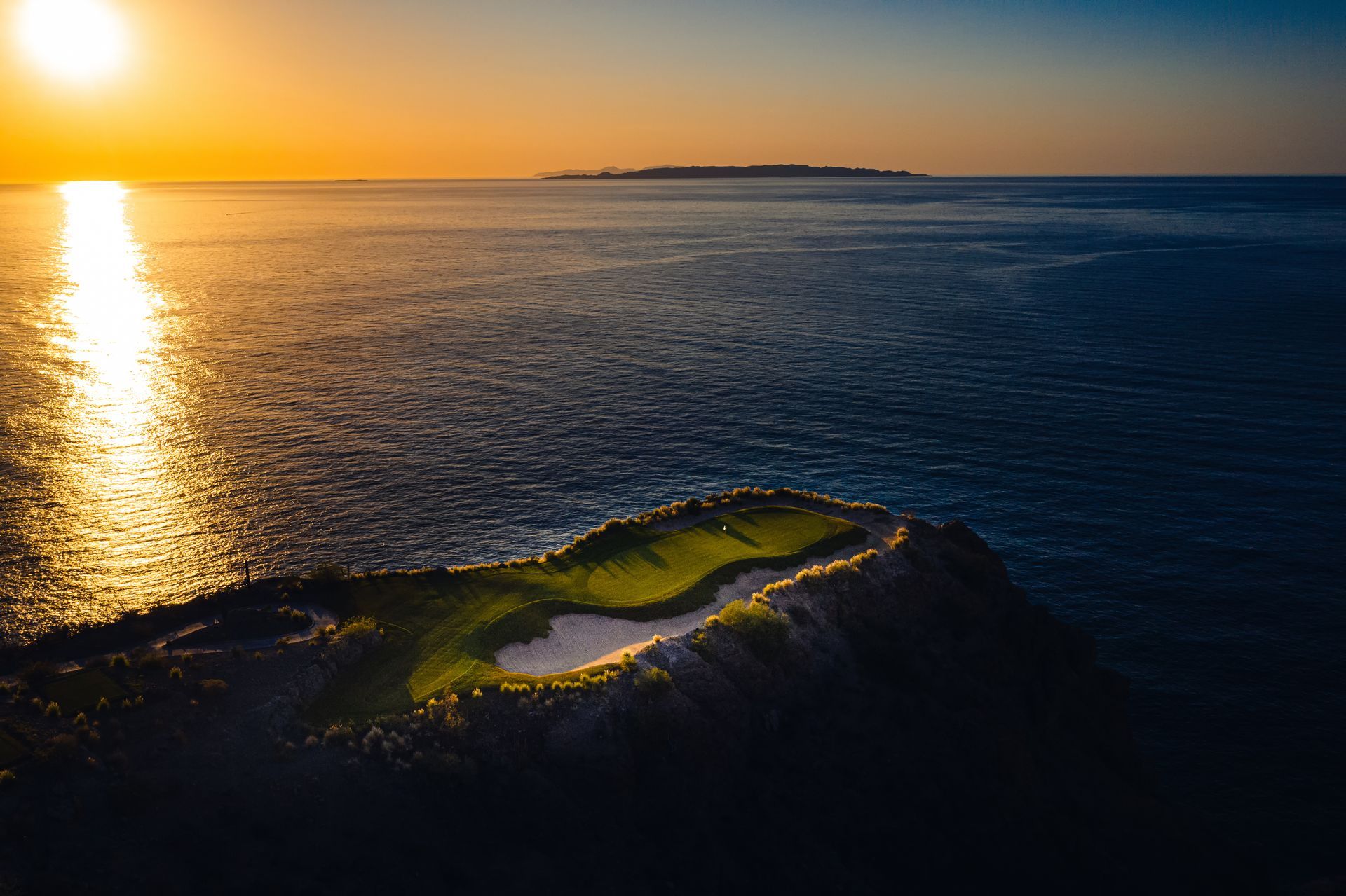 An aerial view of a golf course in the middle of the ocean at sunset.