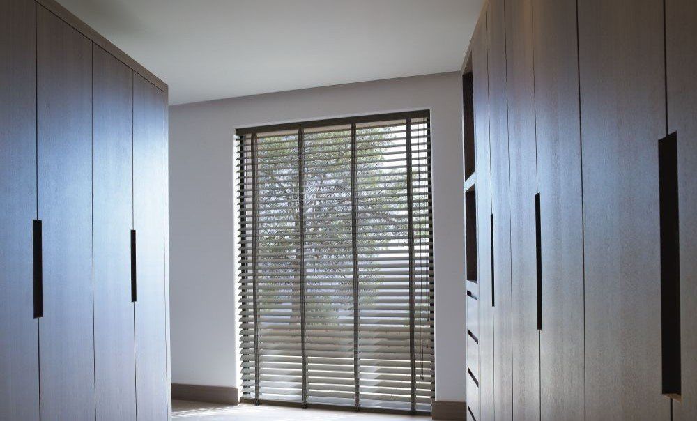 Inloopkast met lichte houten kasten en een raam met horizontale jaloezieën.