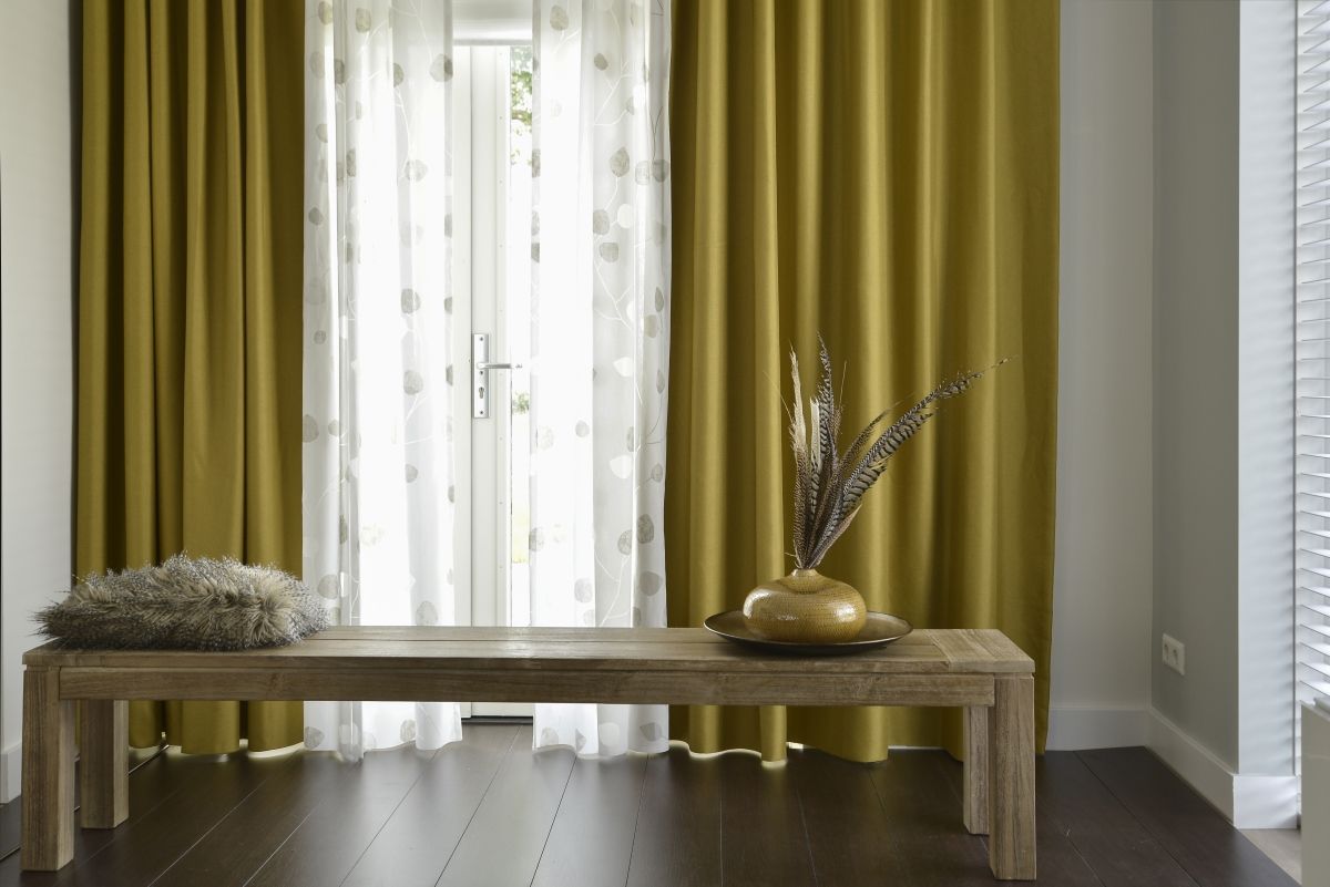 Een houten bank staat voor een raam met gele gordijnen.