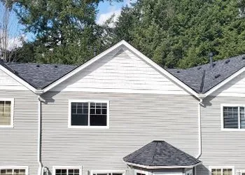Grey multi-unit building with a white gable and a dark roof, set against a backdrop of green trees.