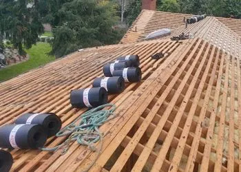 Roof under construction, showing wooden beams, rolls of black roofing material, and rope.