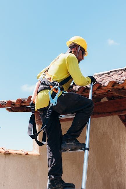 A worker in a yellow shirt, hard hat, and safety harness climbs a ladder toward a tiled roof against a clear blue sky.