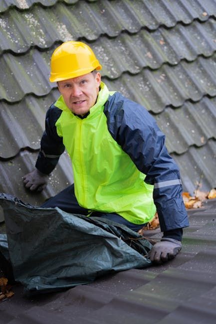 A worker in a yellow hard hat and high-visibility jacket kneeling on a tiled roof while holding a green debris bag.