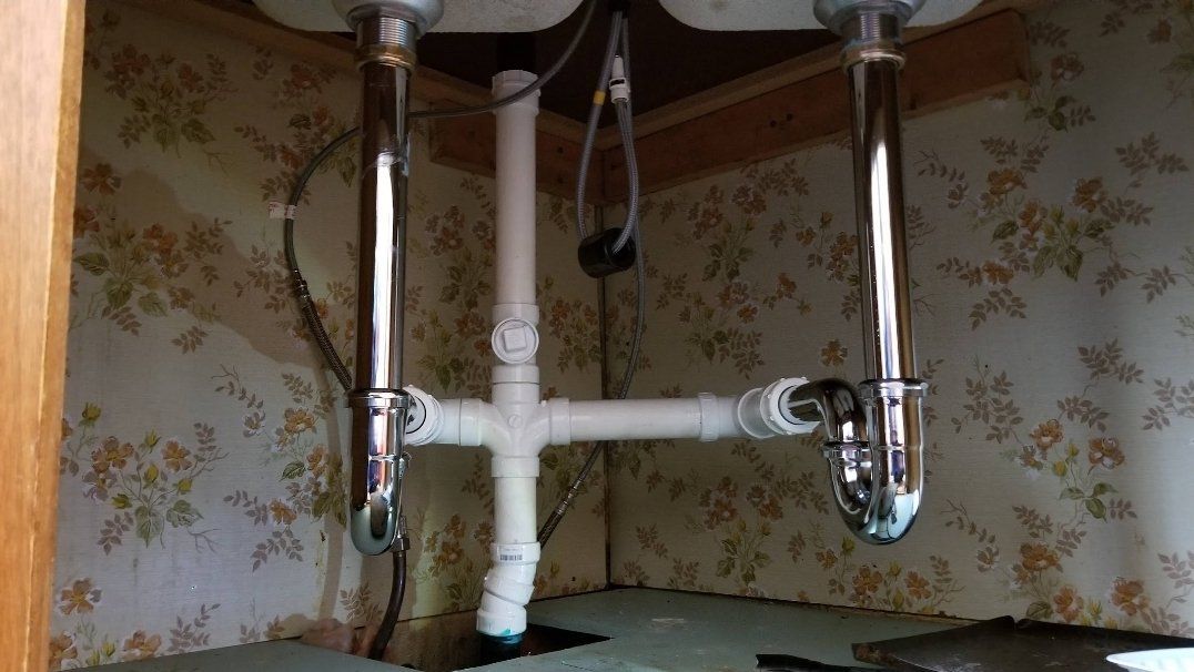 Plumbing pipes under a sink with two chrome drain pipes, white plastic pipes and floral wallpaper.