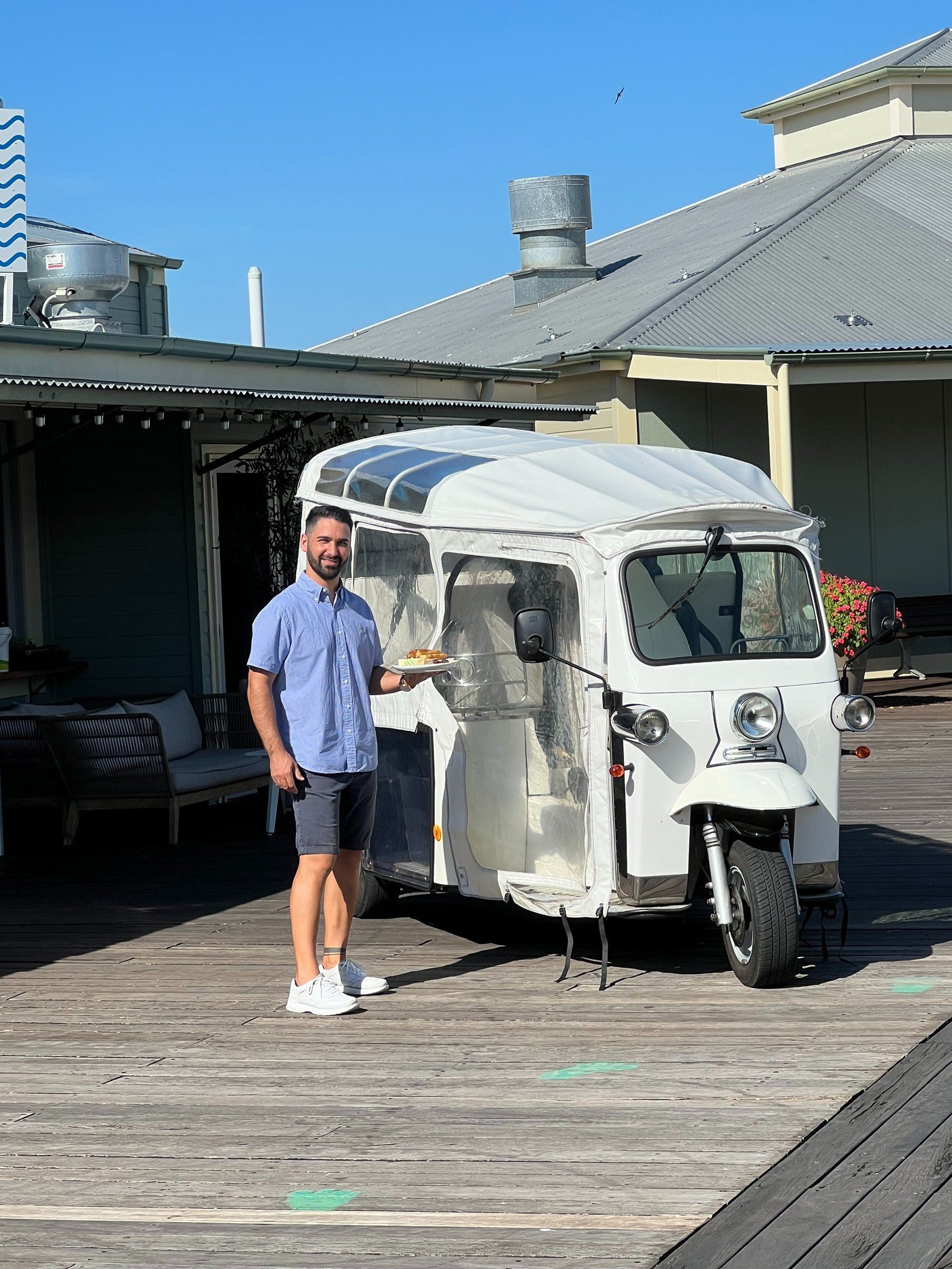 Man beside a white, three-wheeled vehicle with solar panels on a wooden deck; a restaurant in the background.