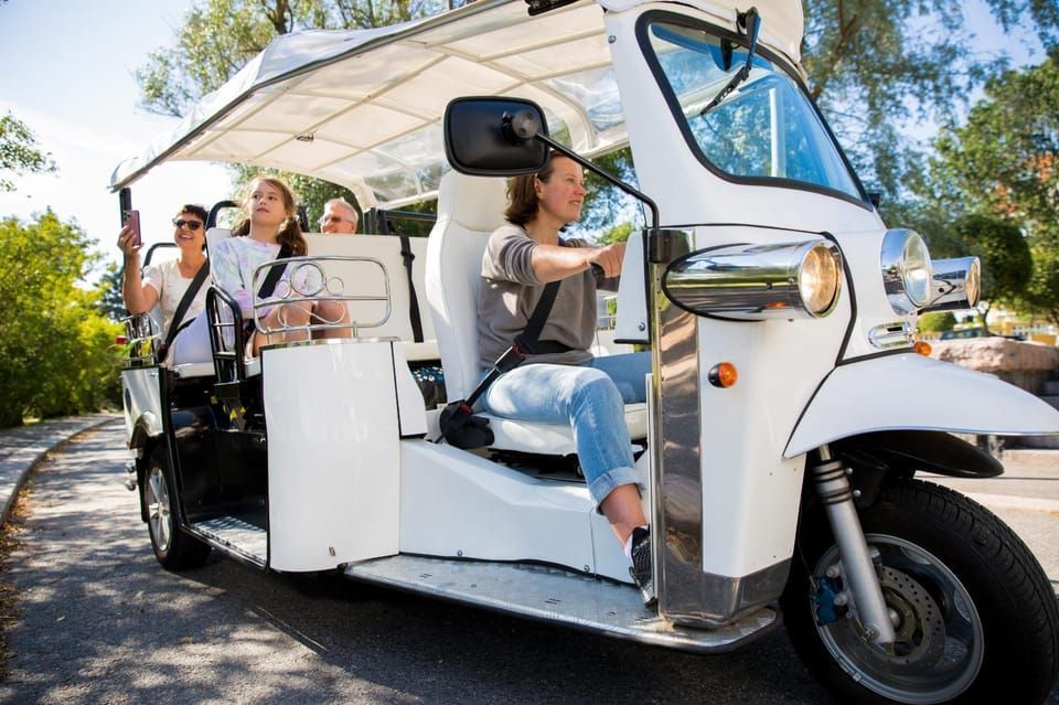White tuk-tuk with passengers driving on a road. Driver and two passengers are visible. Sunny day.