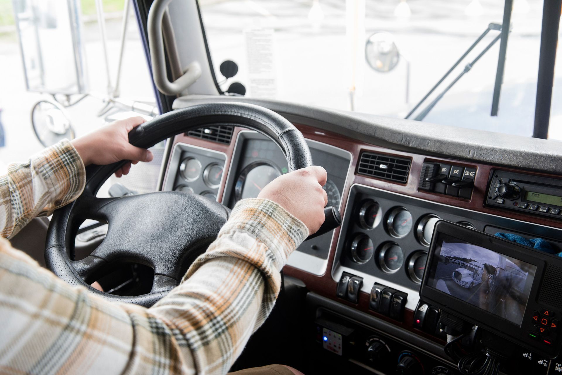 Hands gripping a black steering wheel inside a truck cab. A person wearing a plaid shirt is driving.
