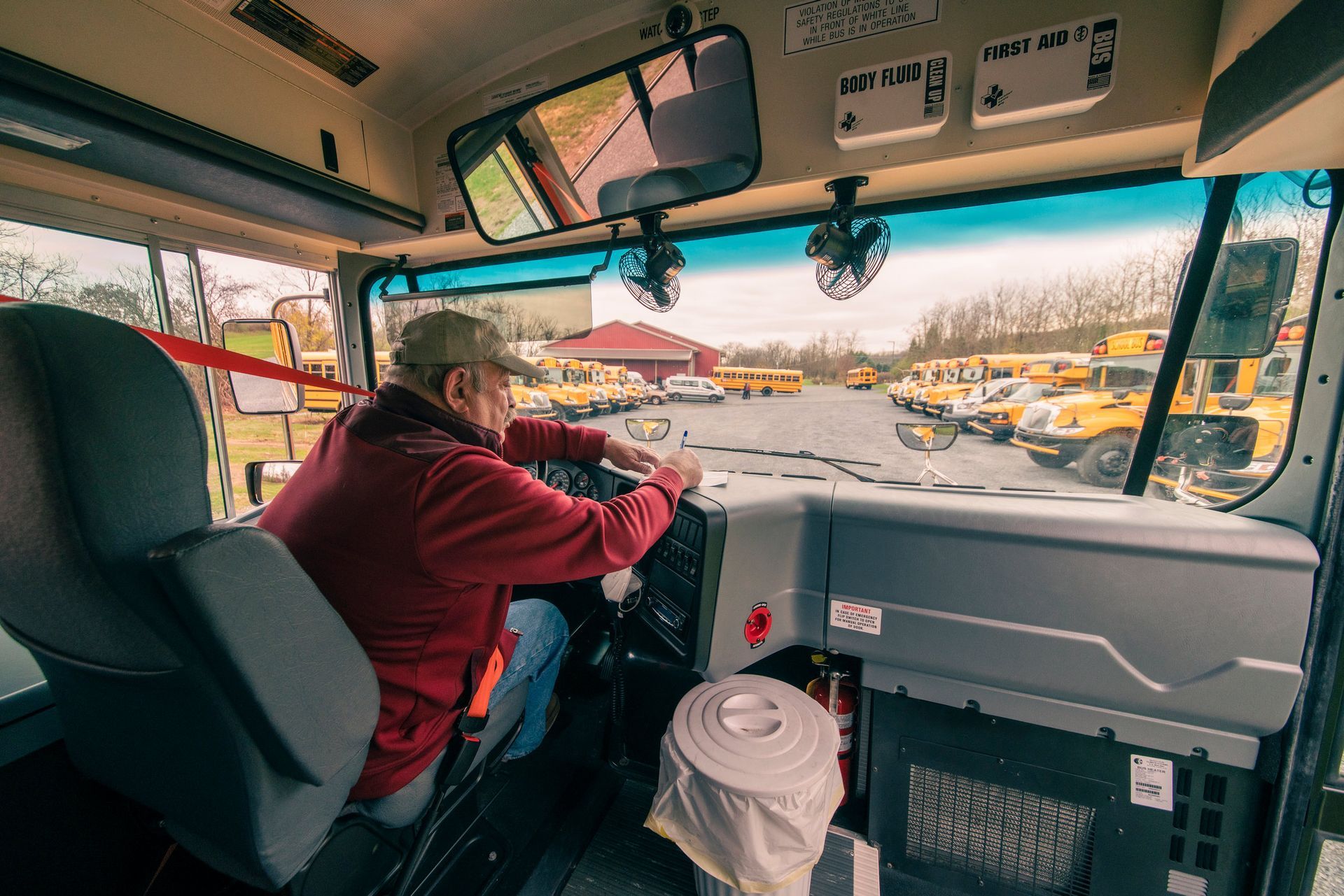 Inside a school bus, a driver in a red jacket operates the steering wheel, looking at the buses in the yard.