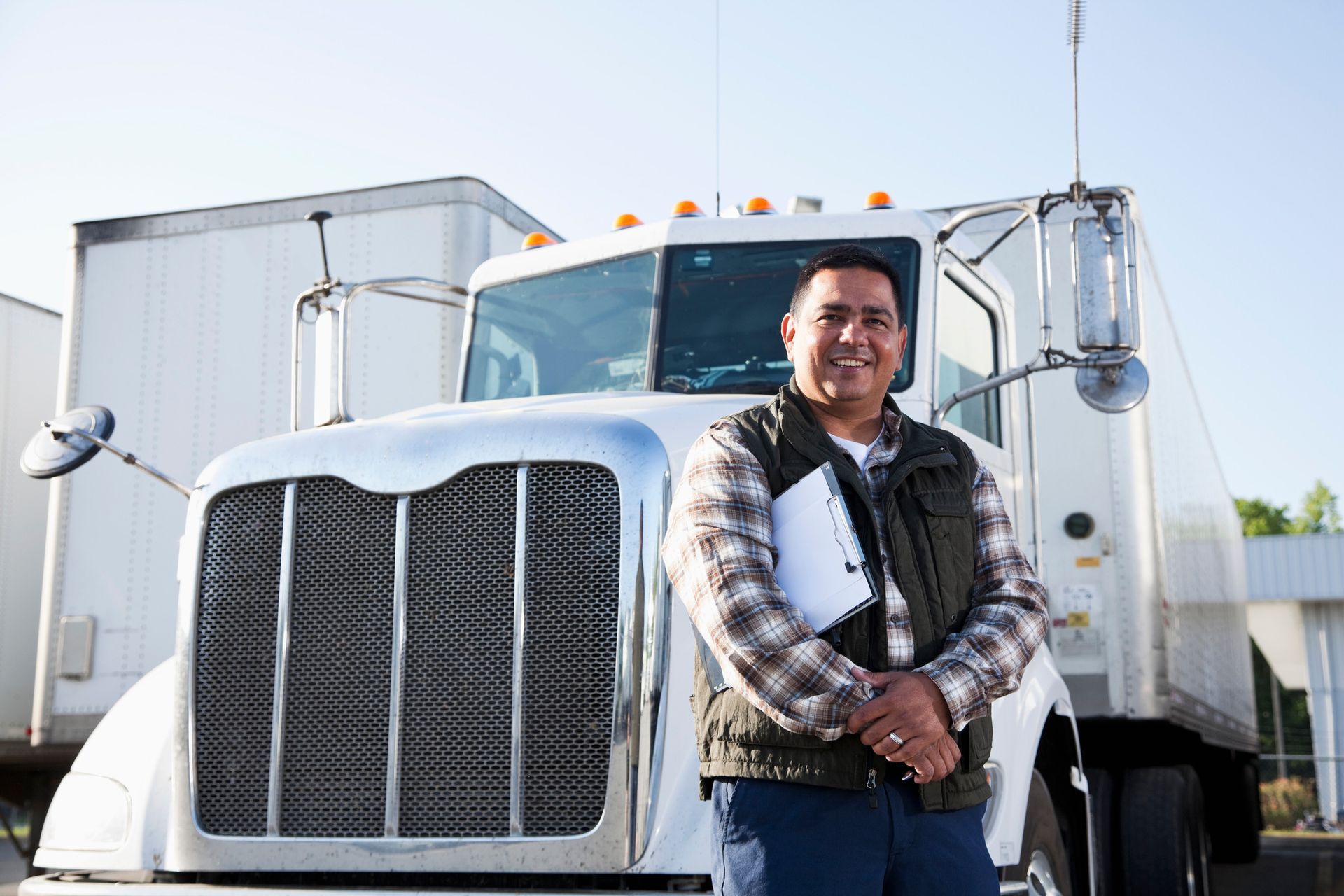 Man in plaid shirt and vest smiles in front of a white semi-truck, holding a tablet.