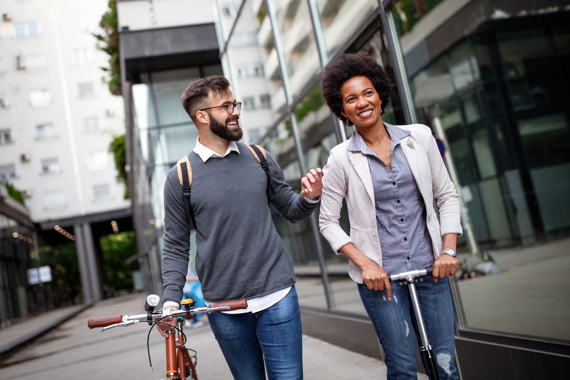 A man and a woman are walking down the street with a bicycle and scooter.