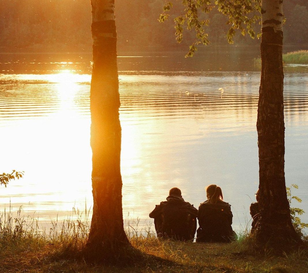 Two figures sit by a lake, silhouetted by the setting sun between two trees.