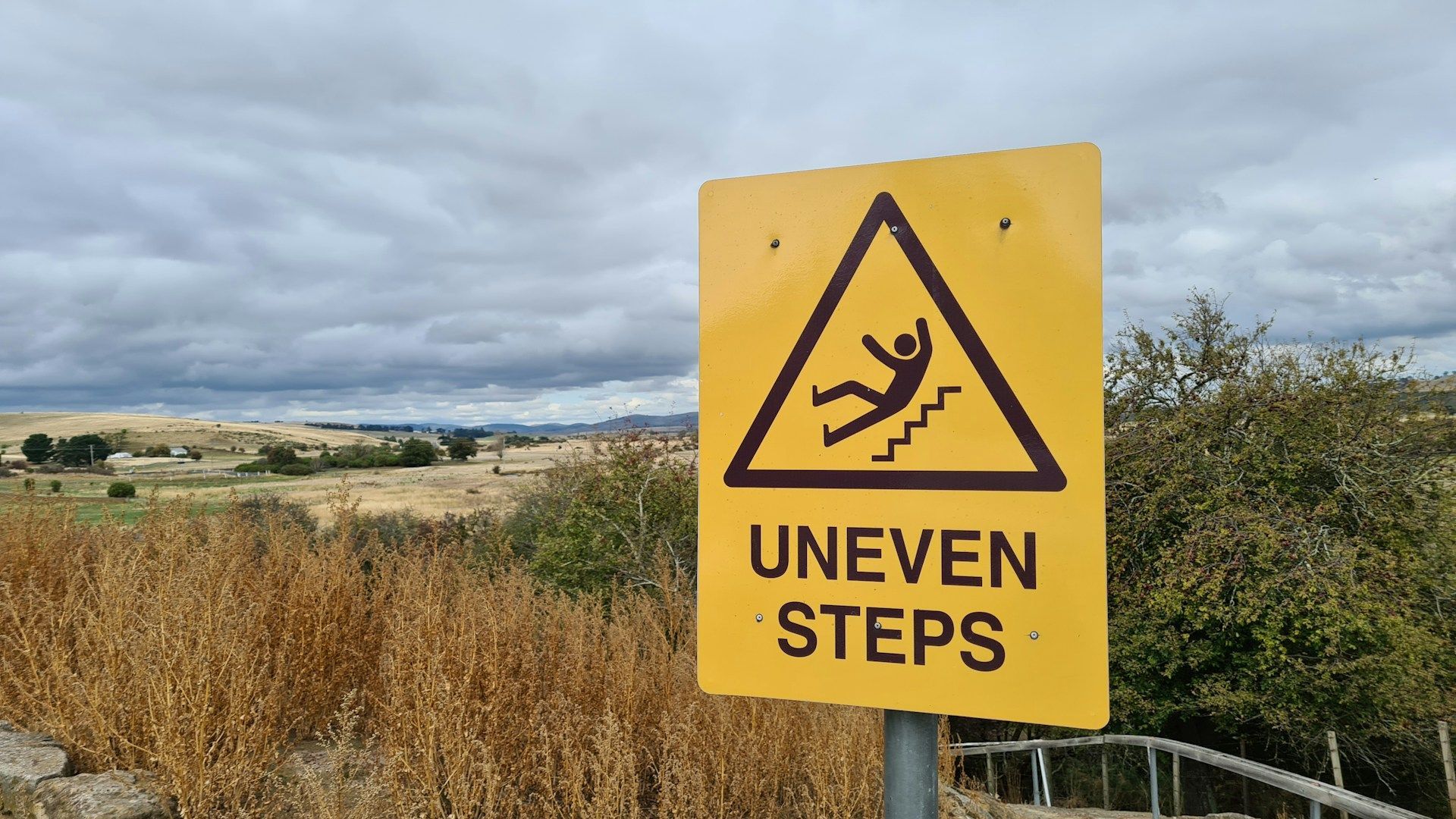 Yellow sign with warning of uneven steps, showing a falling person icon, against a landscape background.