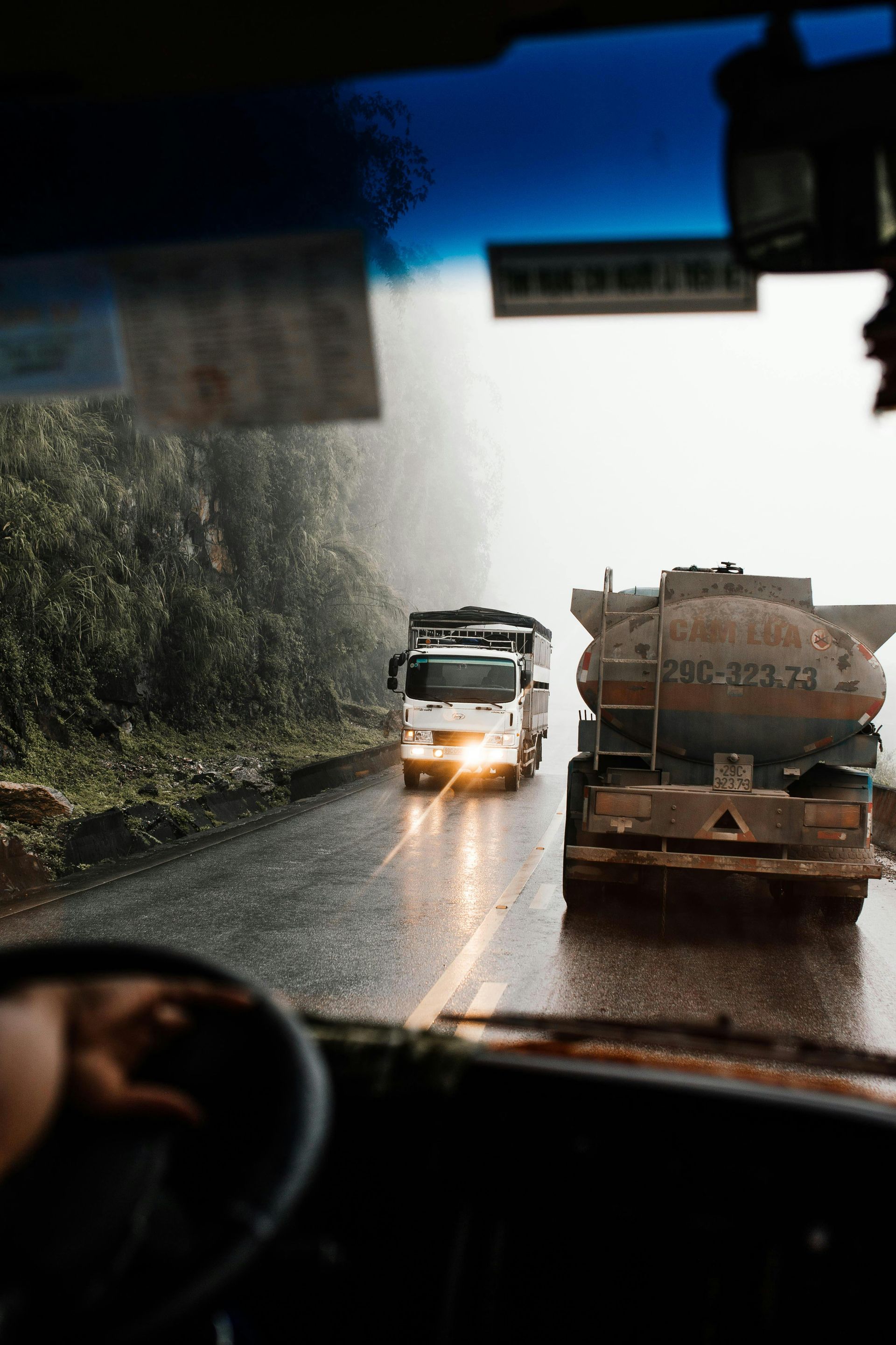 View from inside a vehicle of two trucks driving on a wet road in the rain.