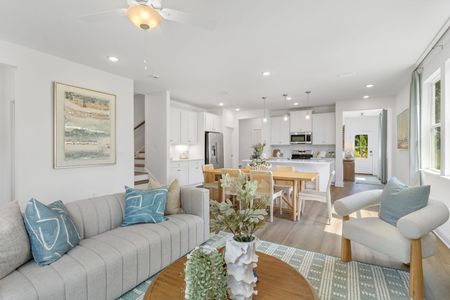 Bright, open living room with light-colored furniture, leading into a kitchen. Soft blues and whites dominate.