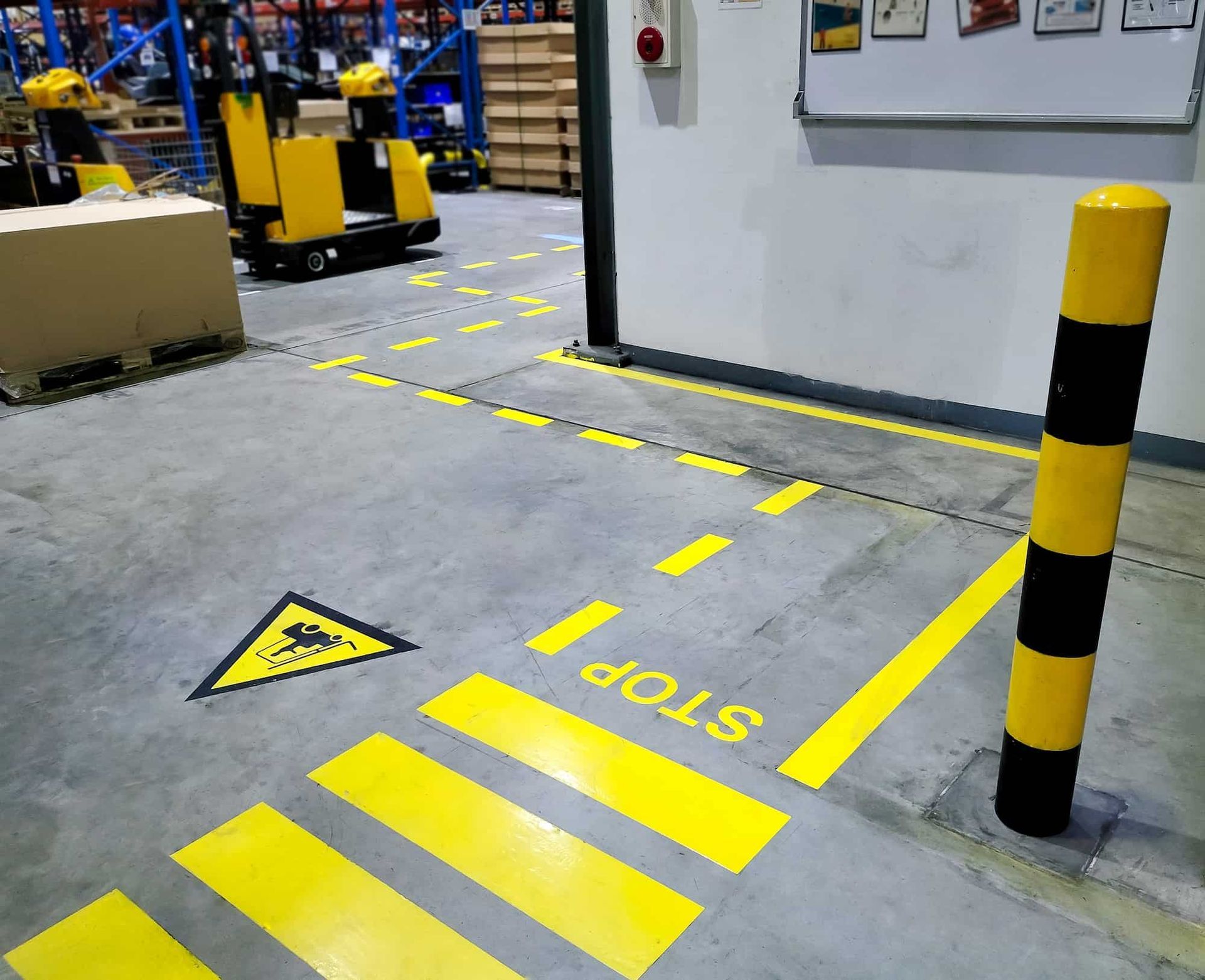 Warehouse floor with yellow safety markings: crosswalk, stop sign, hazard symbol, and bollard.