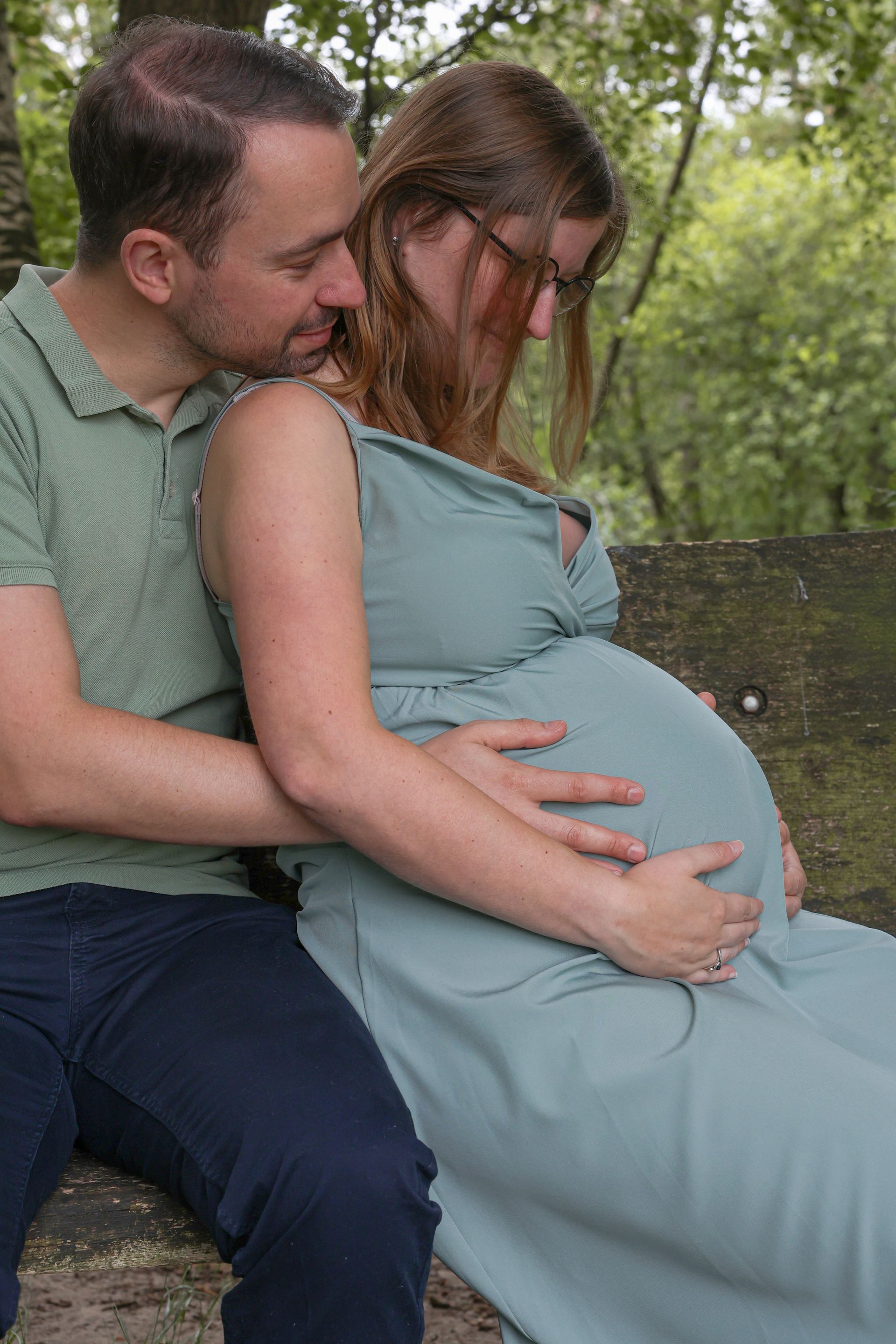 Een stel zit buiten op een bankje; de ​​man omhelst een zwangere vrouw in een blauwe jurk.