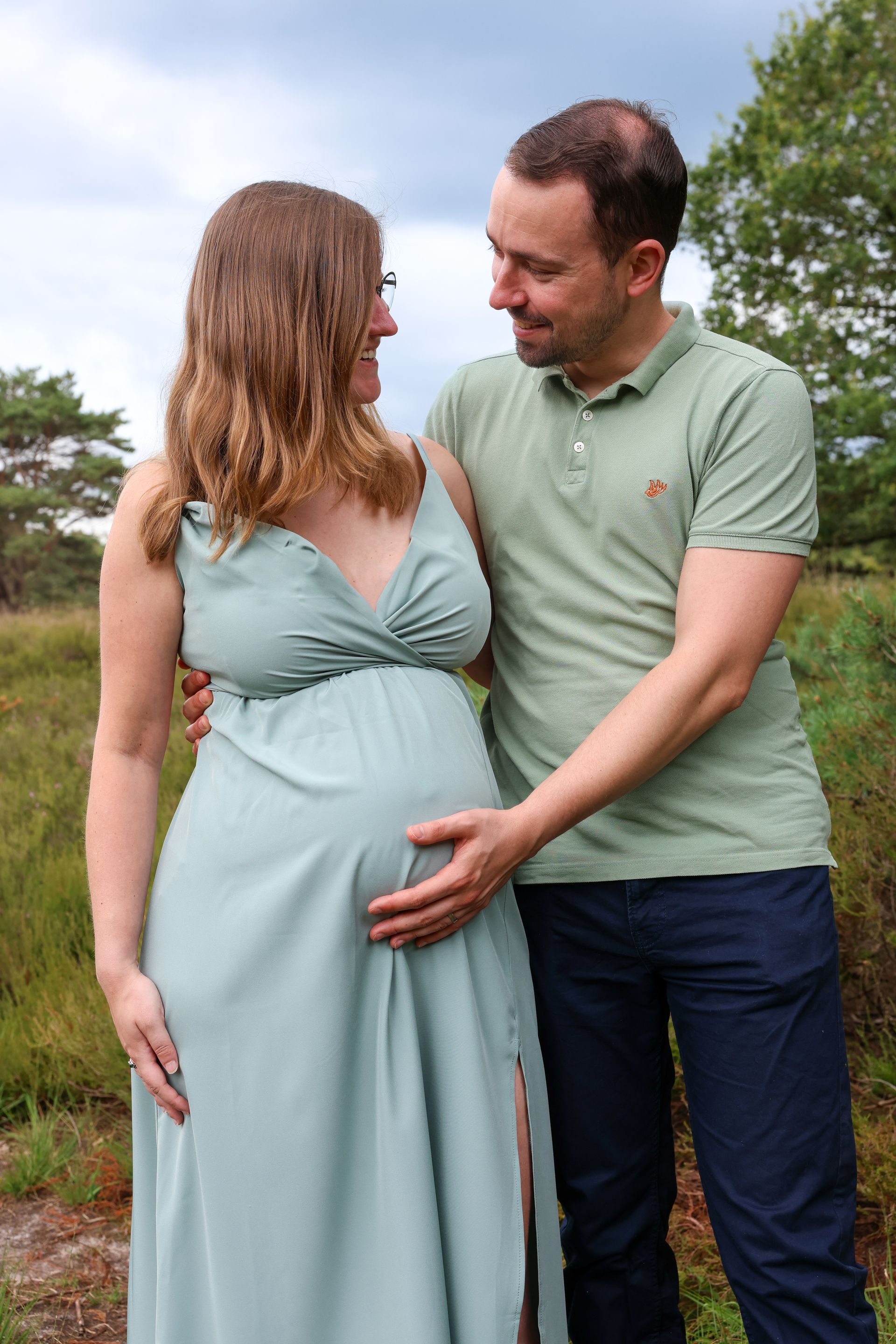 Een zwanger stel staat buiten en lacht terwijl de man de buik van de vrouw vasthoudt.