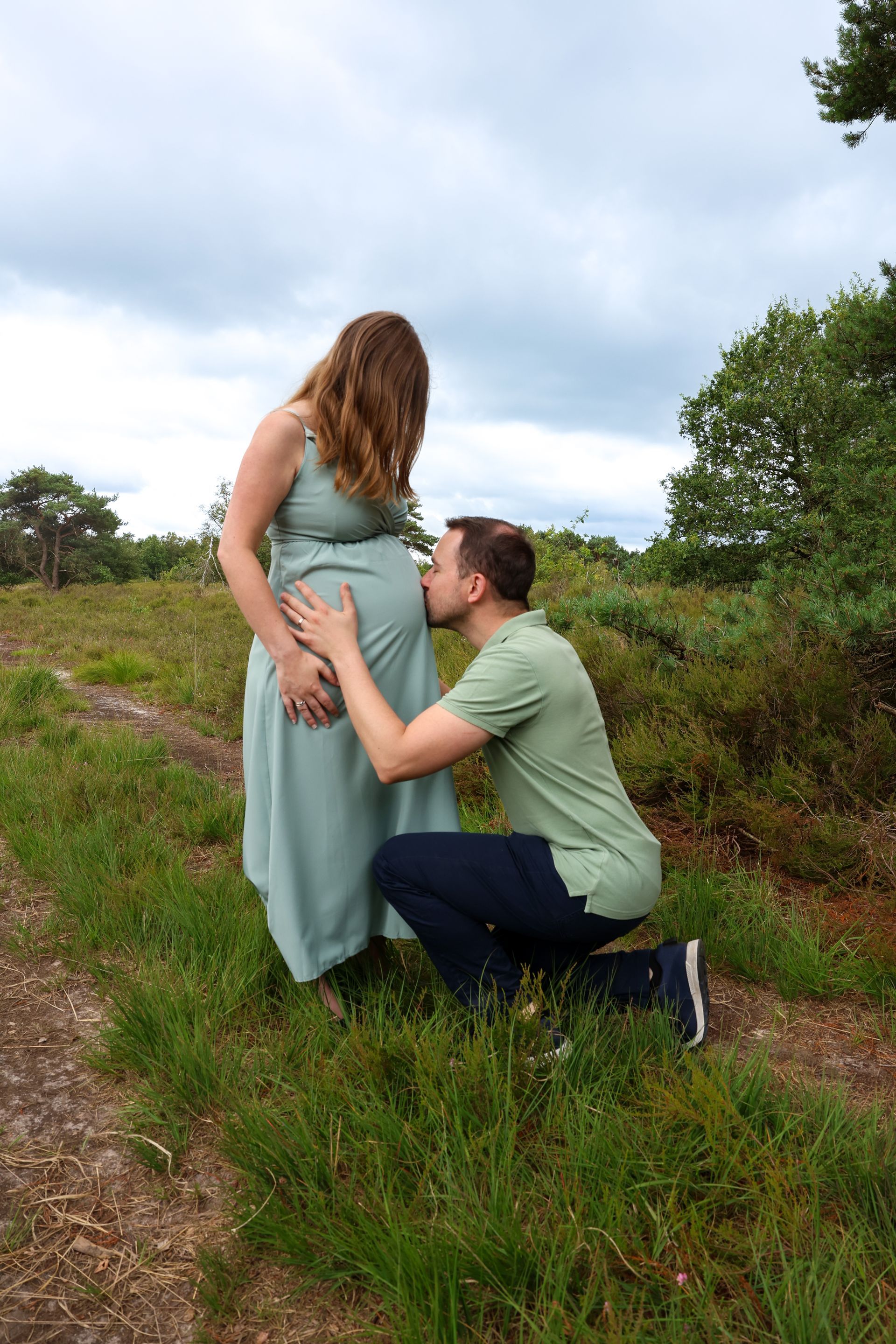 Een man knielt neer en omhelst een zwangere vrouw in een groene jurk in een grasveld onder een bewolkte hemel.