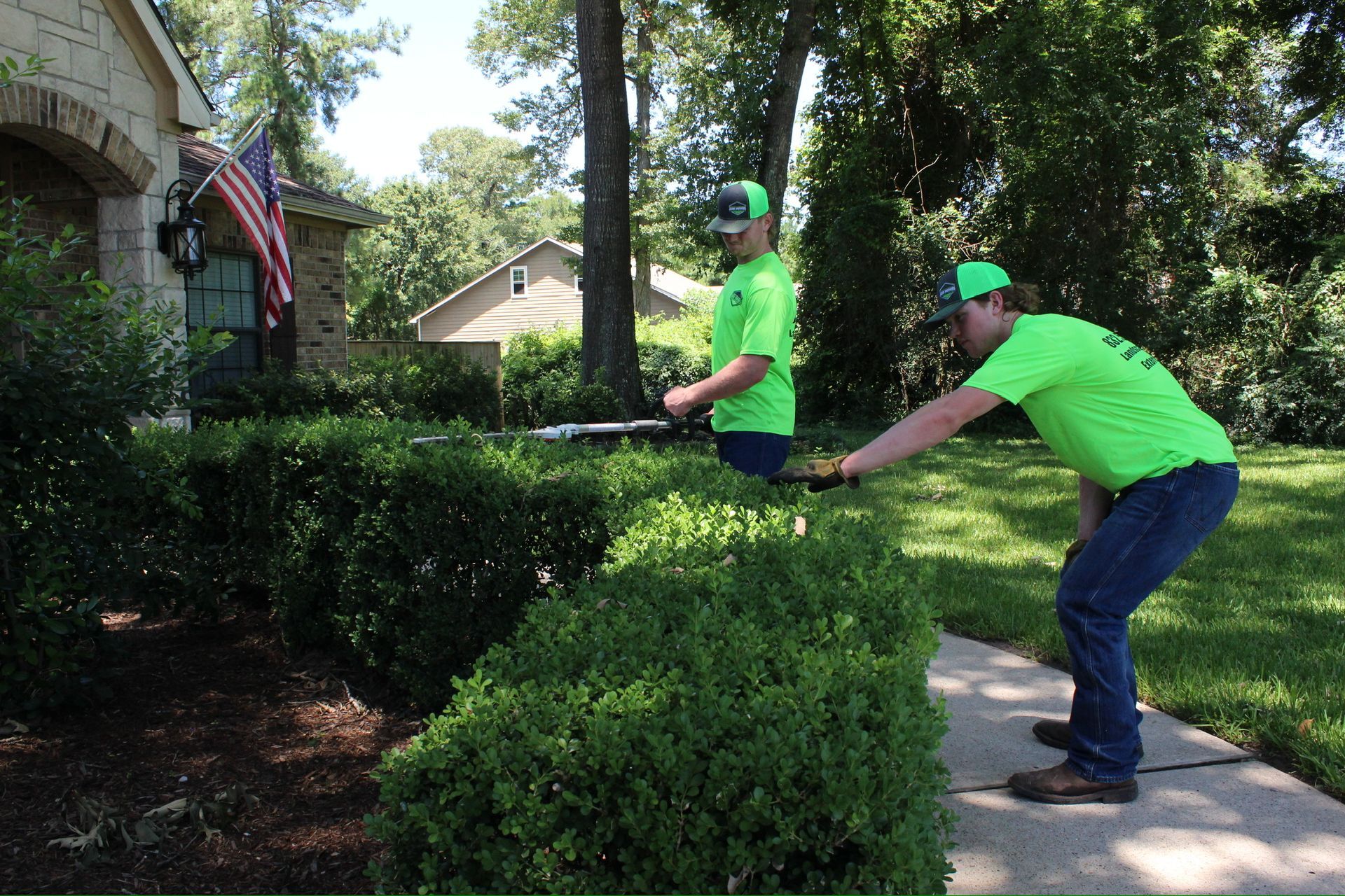 Two men in green shirts are cutting bushes in front of a house.