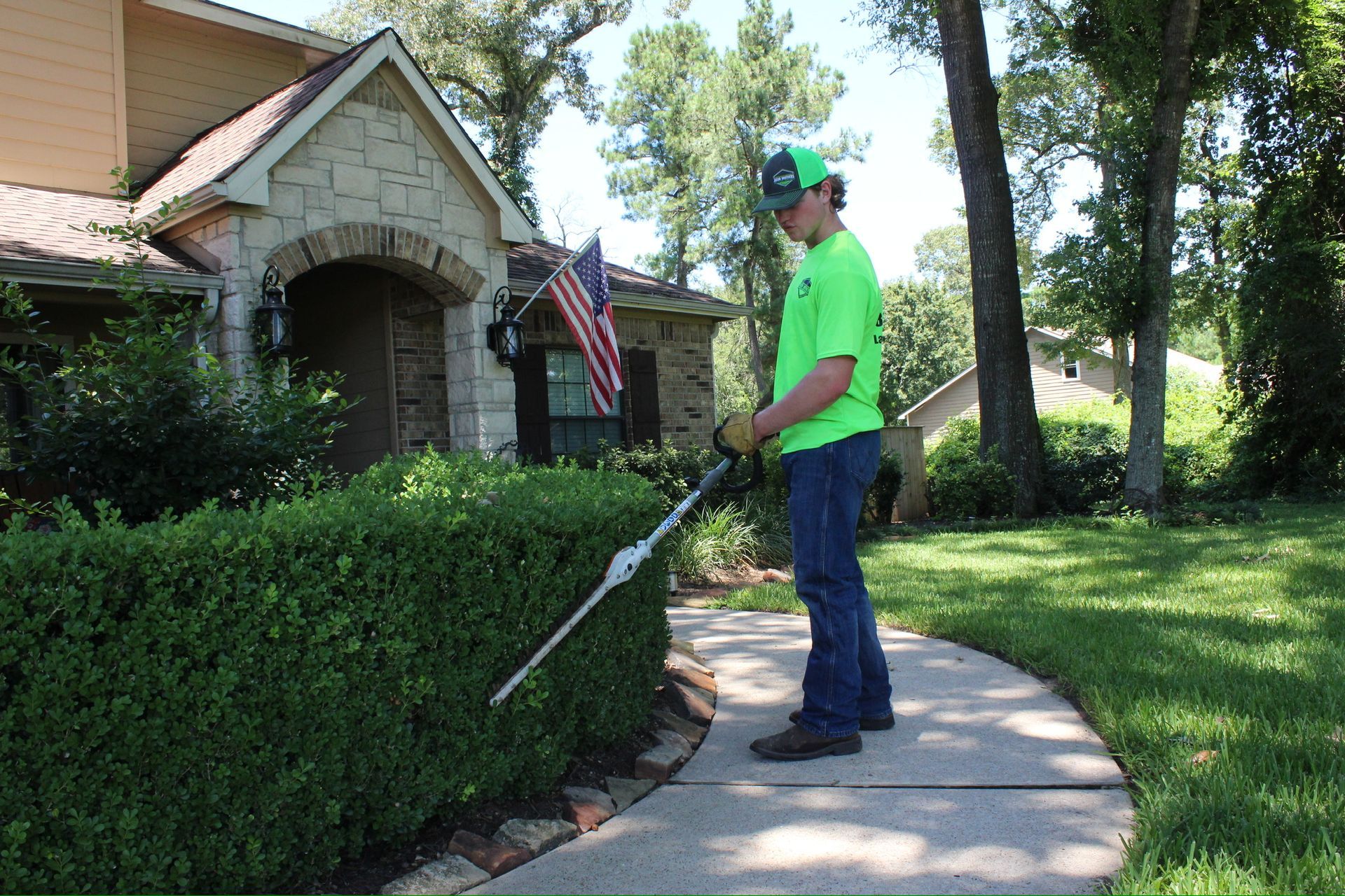 A man is standing on a sidewalk cutting a hedge with a lawn mower.