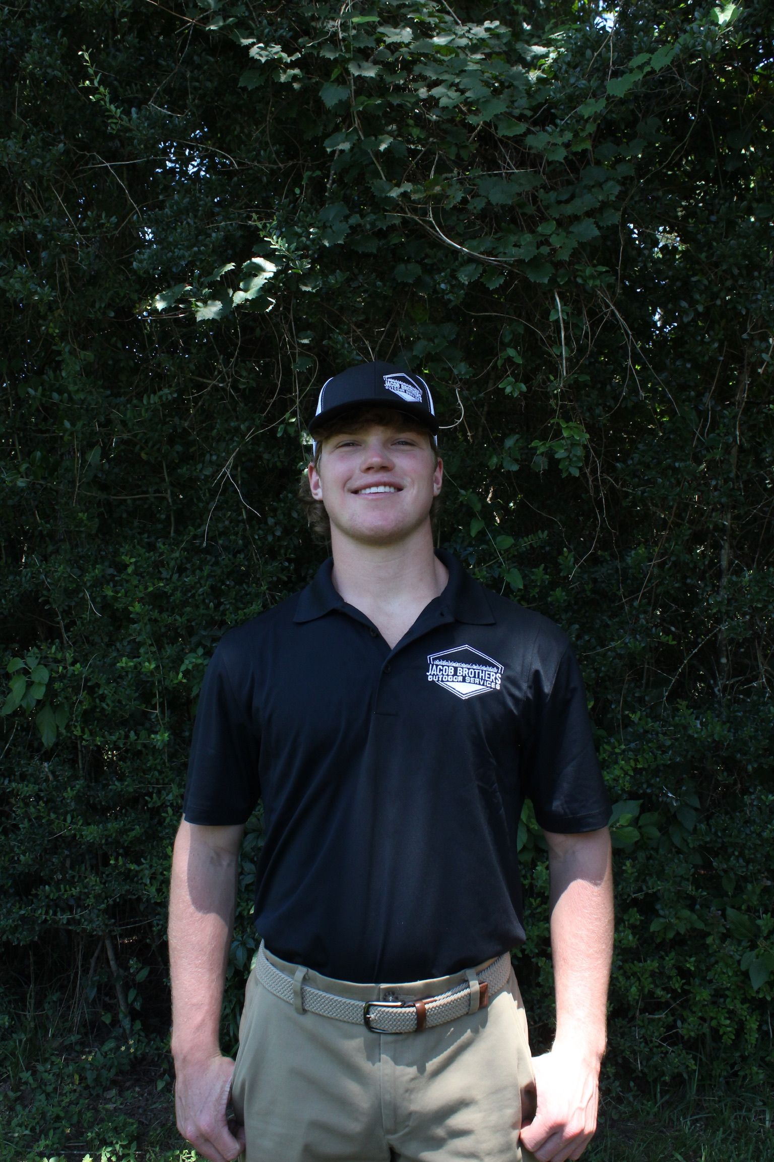 A young man wearing a black shirt and a hat is standing in front of trees.