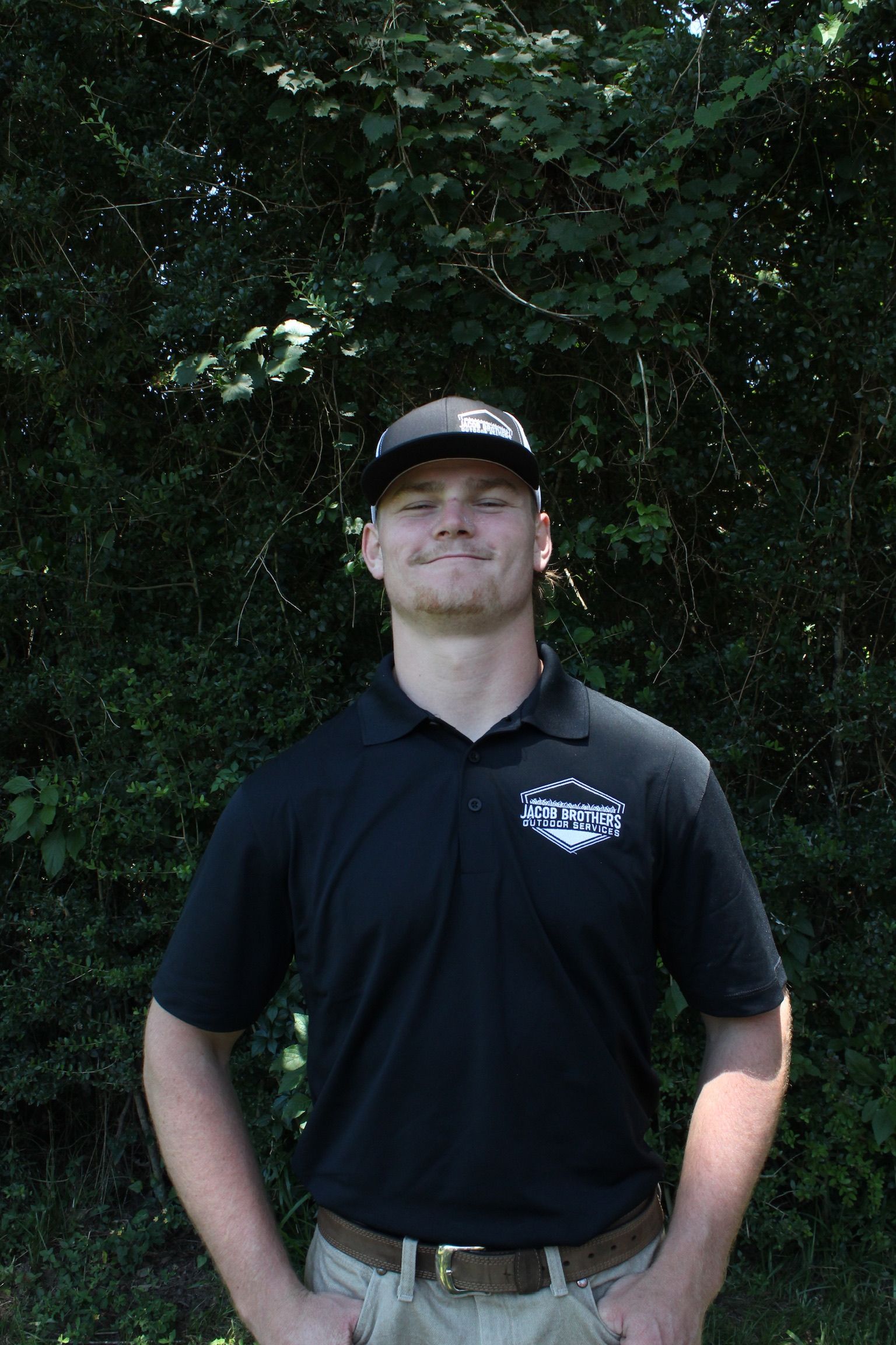 A man wearing a black polo shirt and a hat is standing in front of trees.