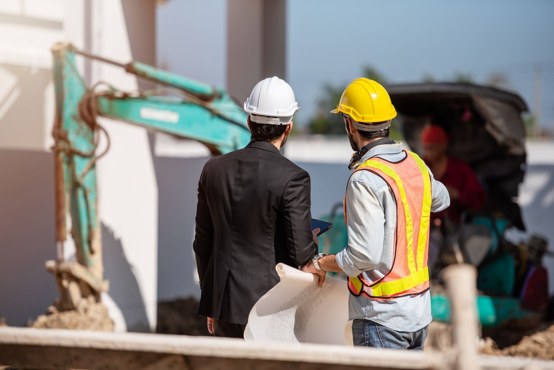 Two construction workers in hard hats reviewing work near an excavator at a building site