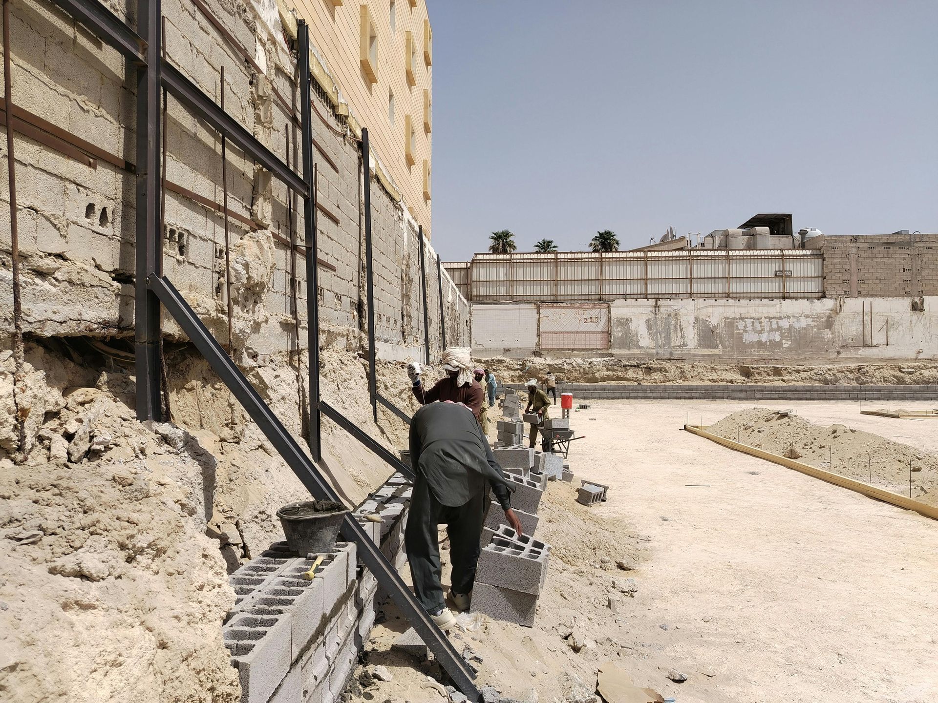 Two people walk along a gravel path beside a high stone wall and metal railings in a sunlit ruin.