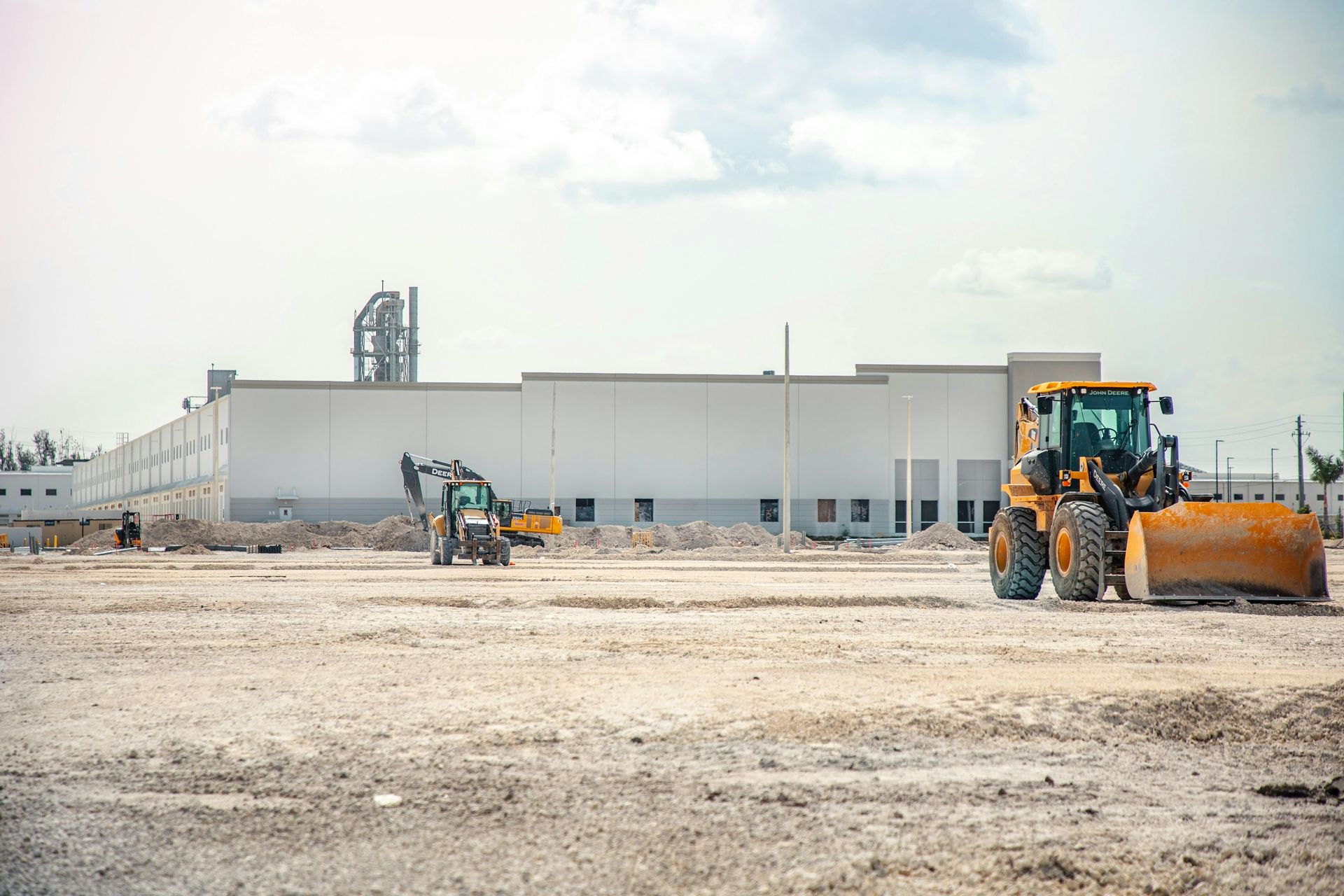 Construction site with a white industrial building and two yellow earthmoving machines on a gravel lot
